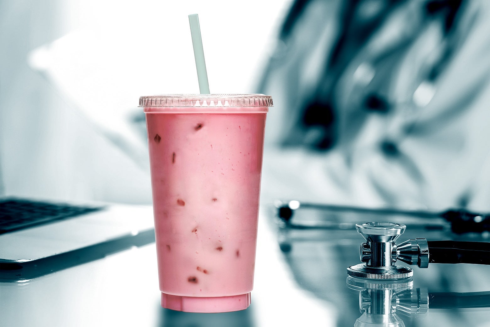 A smoothie next to a laptop and a stethoscope on a table, with a doctor looking at a chart in the background