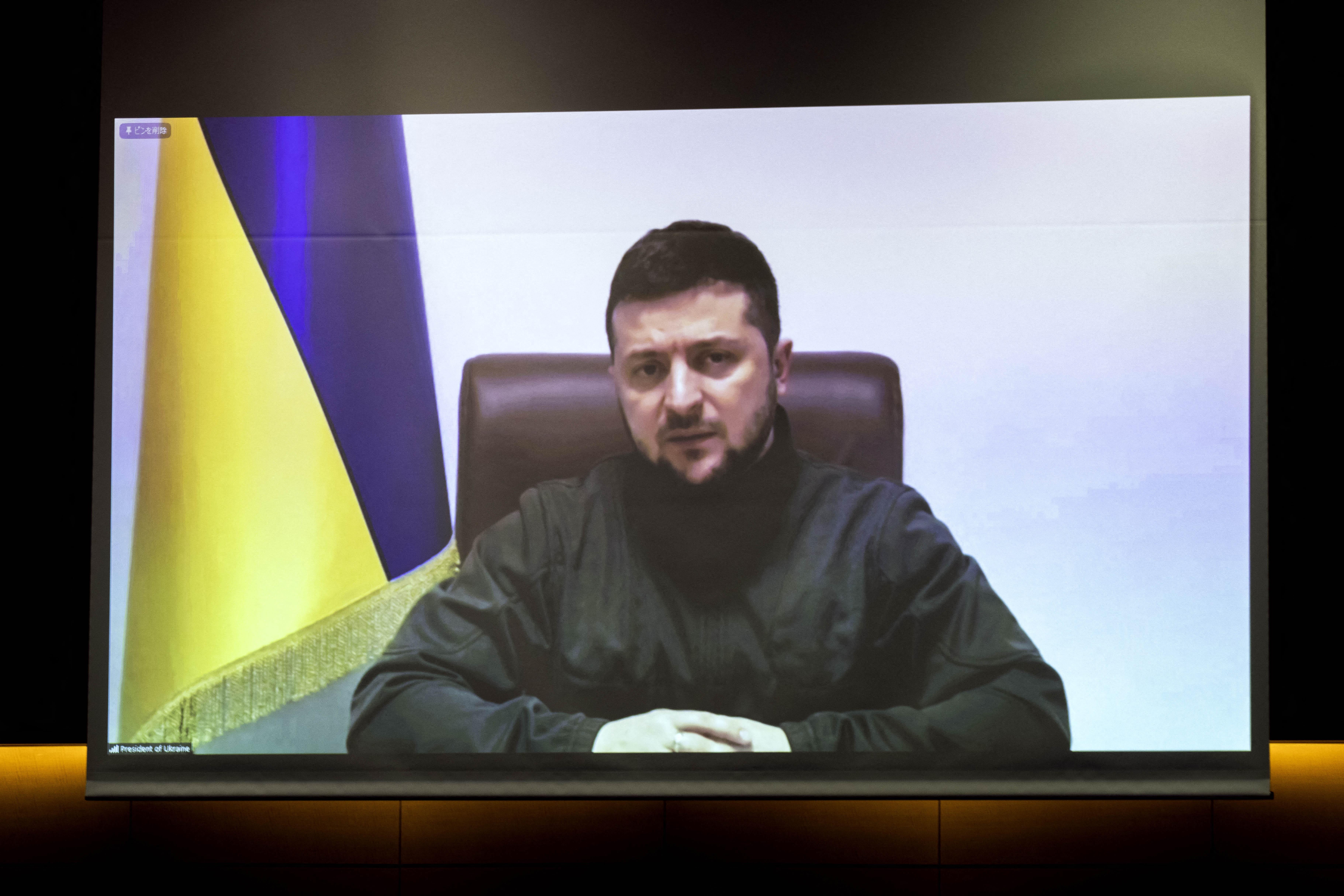 A man on a video screen sits at a table in front of the Ukrainian flag.