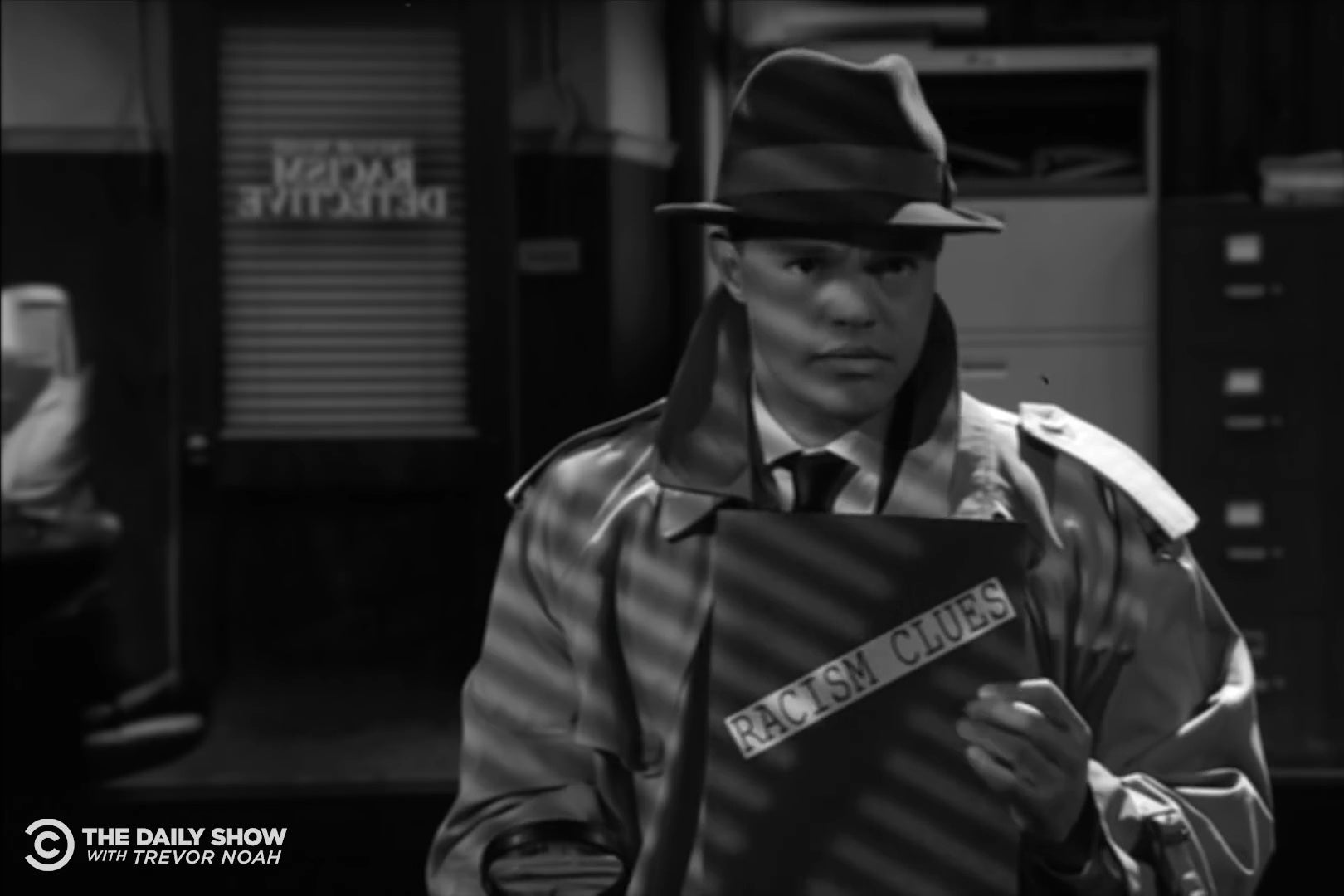 Trevor Noah dressed as a 1940s detective, holding a folder labeled "racism clues."