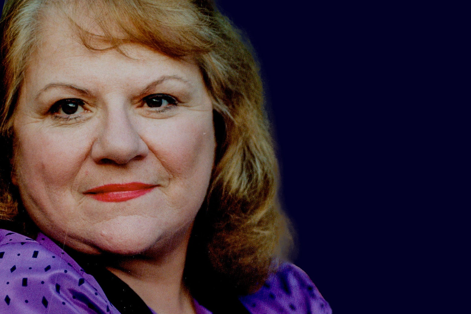 A middle-aged woman with shoulder-length reddish hair, red lipstick, and a purple top stares straight ahead.