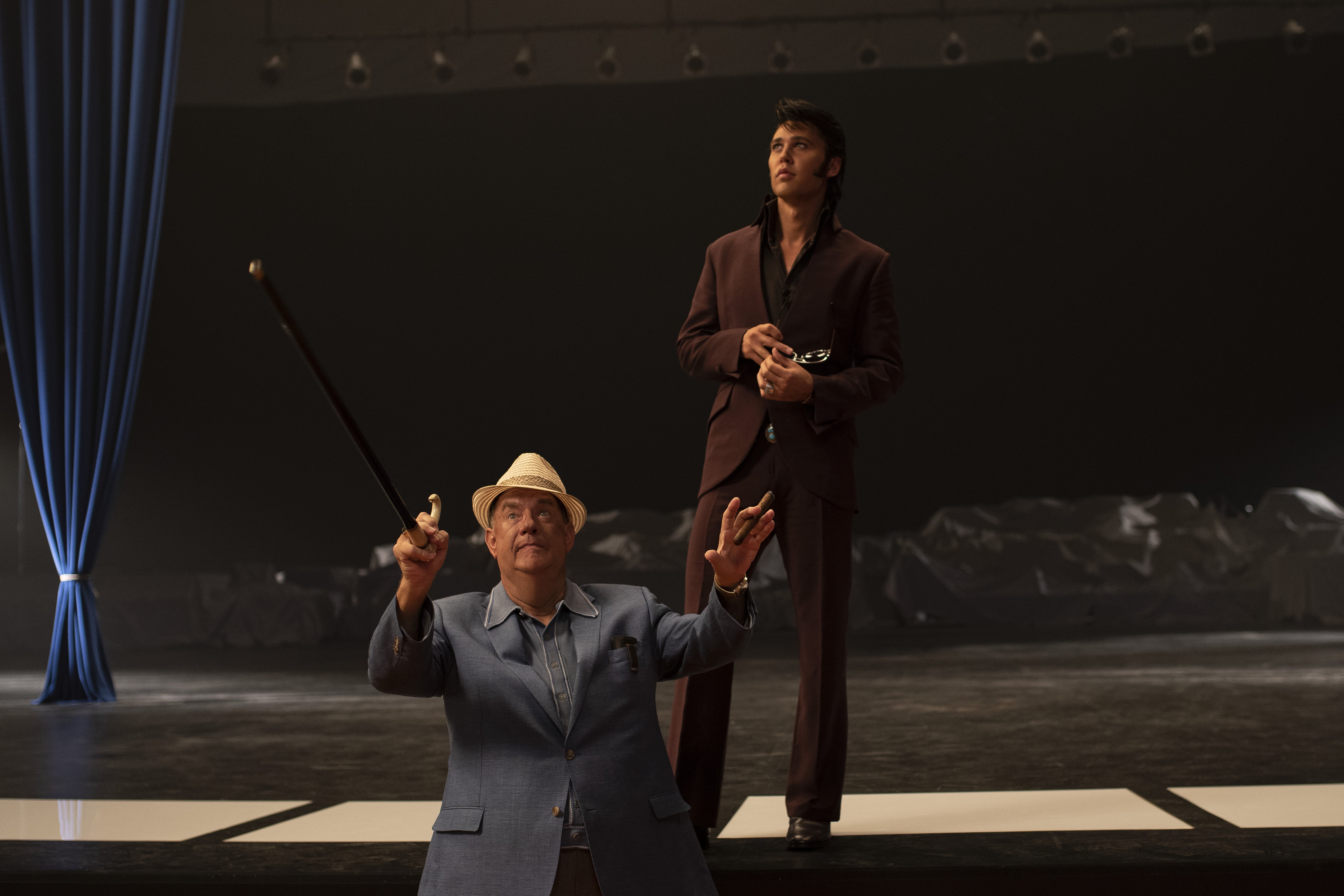 Hanks, in a fat suit under a regular, powder-blue suit, holds up his hands in front of a stage, as if presenting a grand vision, with a hat on his head and a cane in one hand. Behind him, a handsome Austin Butler, tanned and standing on the empty page in a burgundy suit, appears to consider the vision while holding his signature sunglasses in one hand.