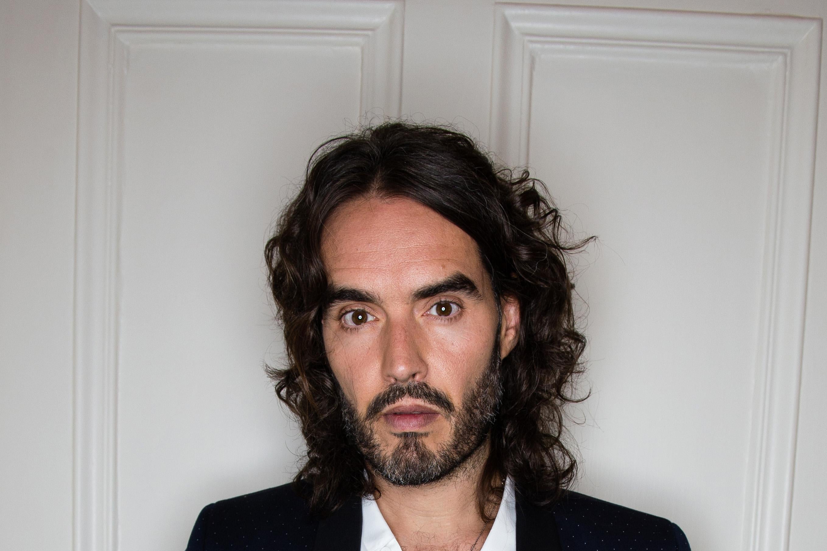 Russell Brand stands before a white wall.