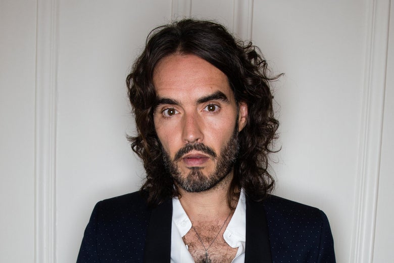 Russell Brand stands before a white wall.