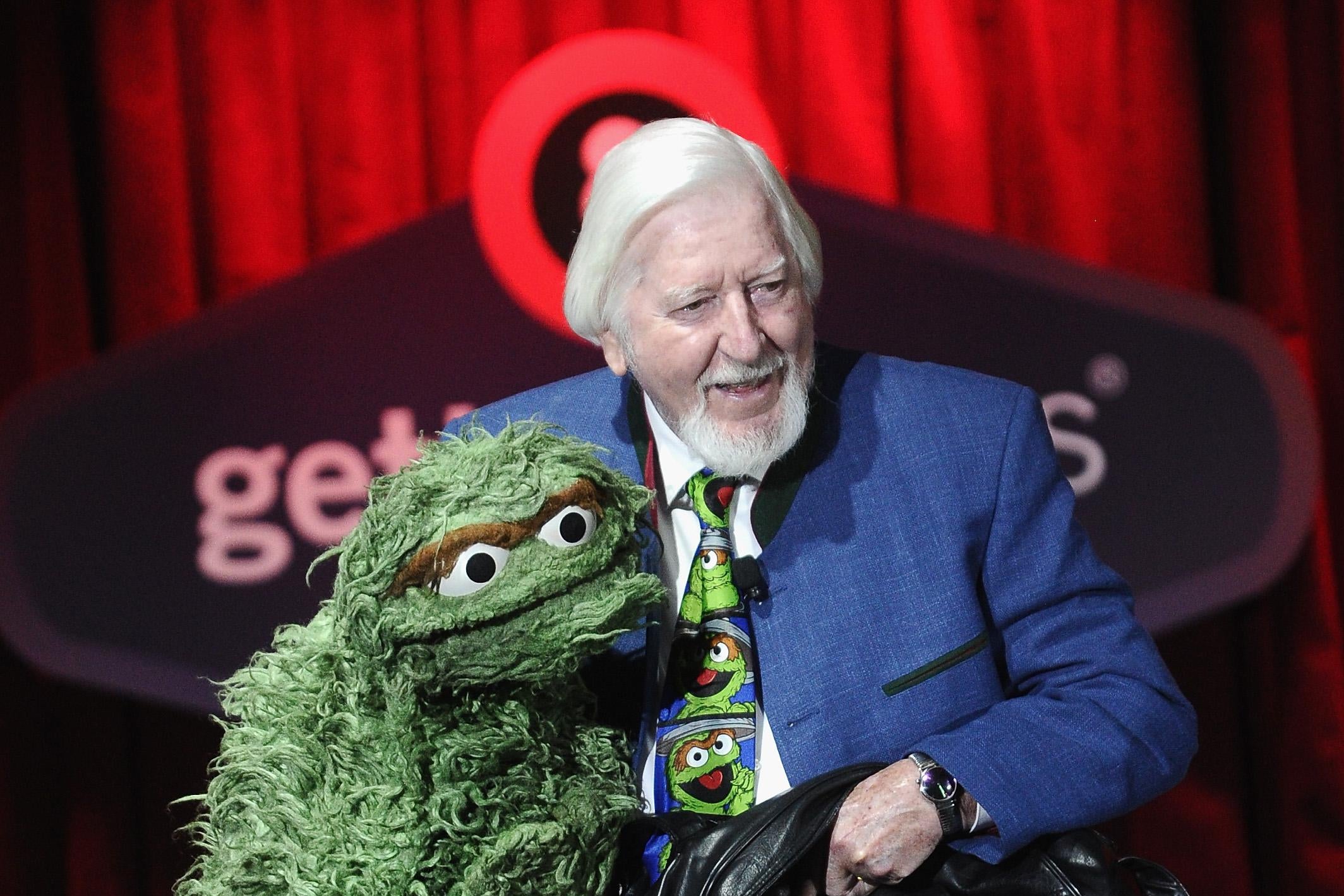 Caroll Spinney, with long white hair and a white beard, sits on a stage operating a green, fuzzy Oscar the Grouch puppet. 
