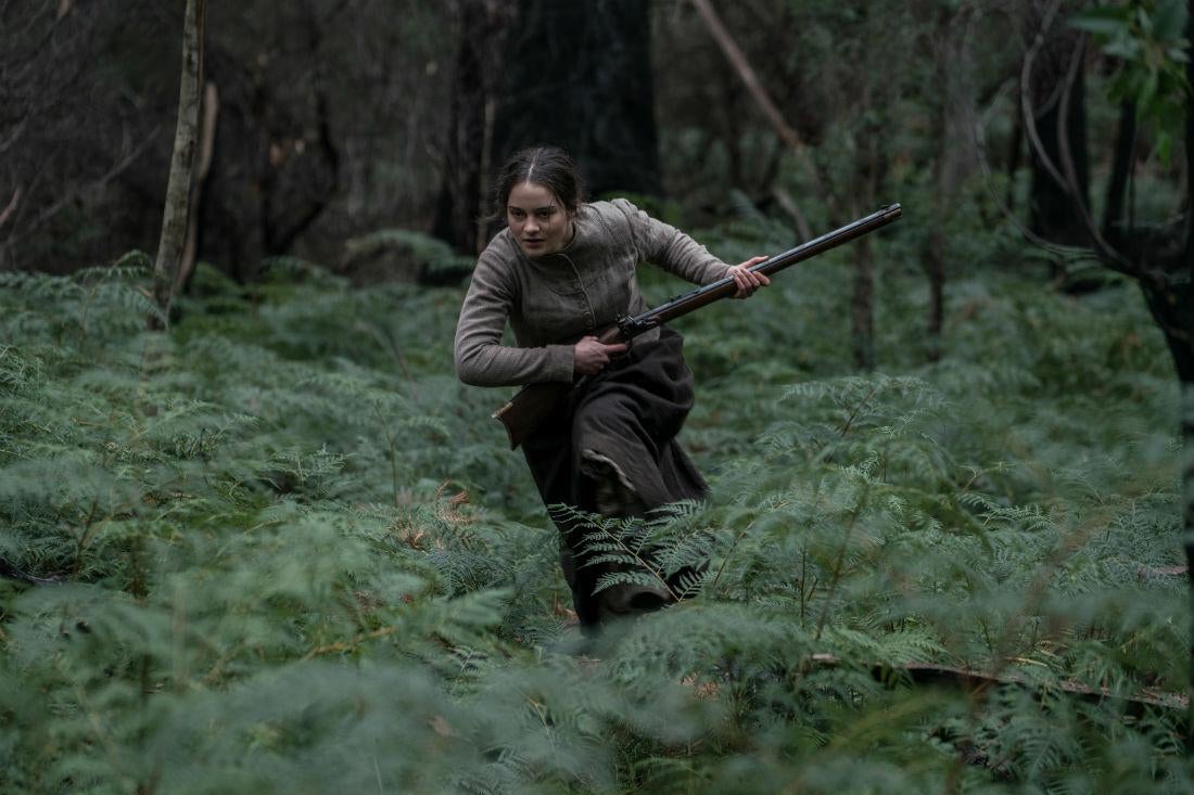 A young woman in a colonial-era peasant dress races through the woods holding a musket.
