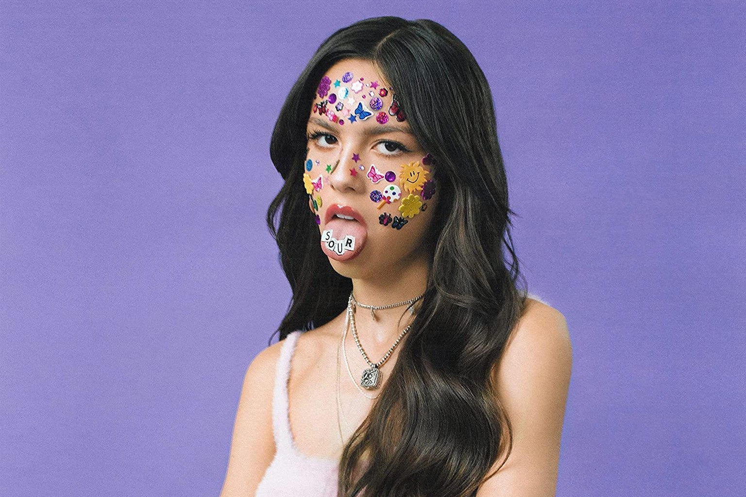 A woman with long brown hair and a white tank top stares at the camera with her tongue out. She has multicolored stickers all over her face and stickers that spell out the word SOUR on her tongue.