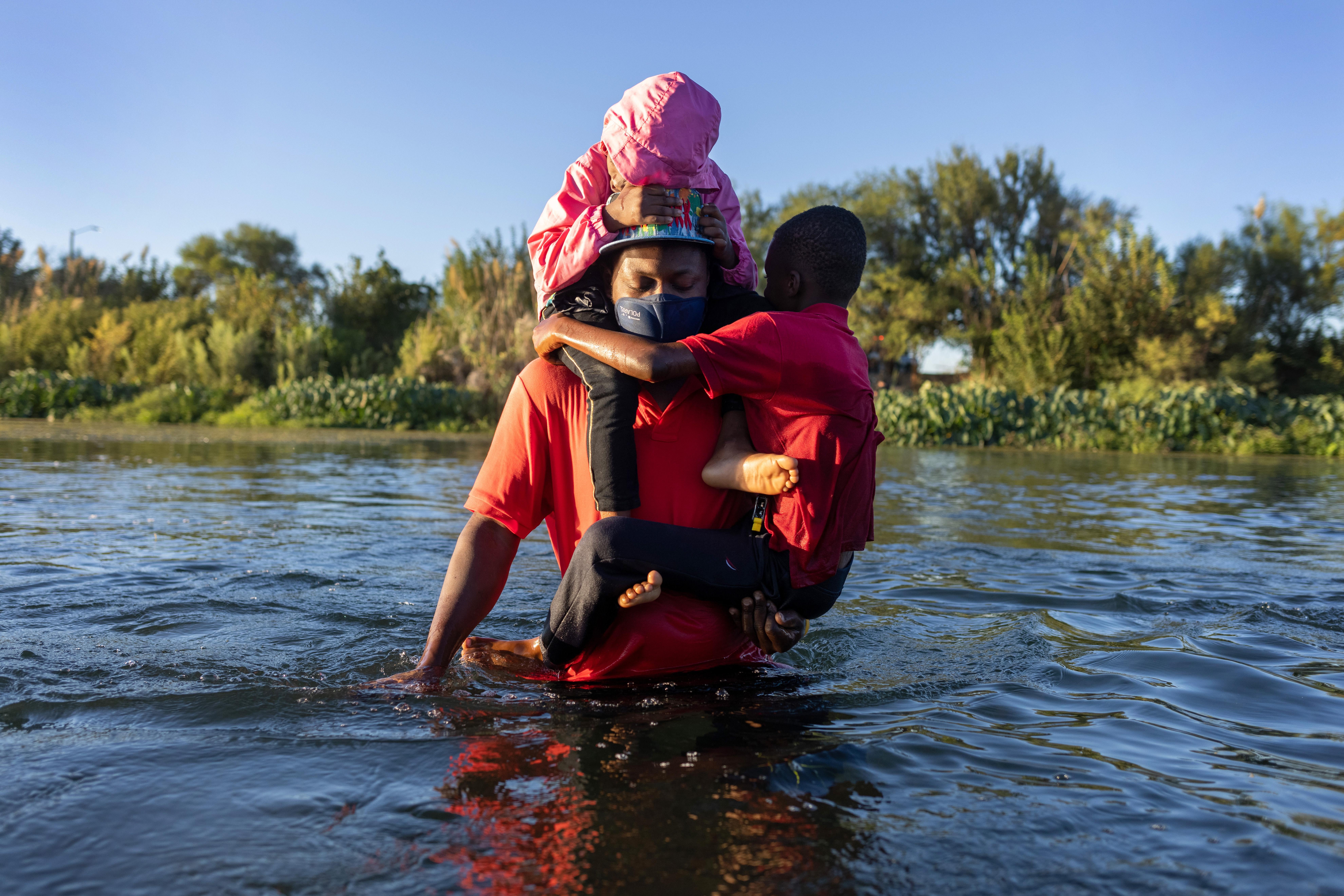 Two small children wrapped around their father's back and torso as he wades through a river
