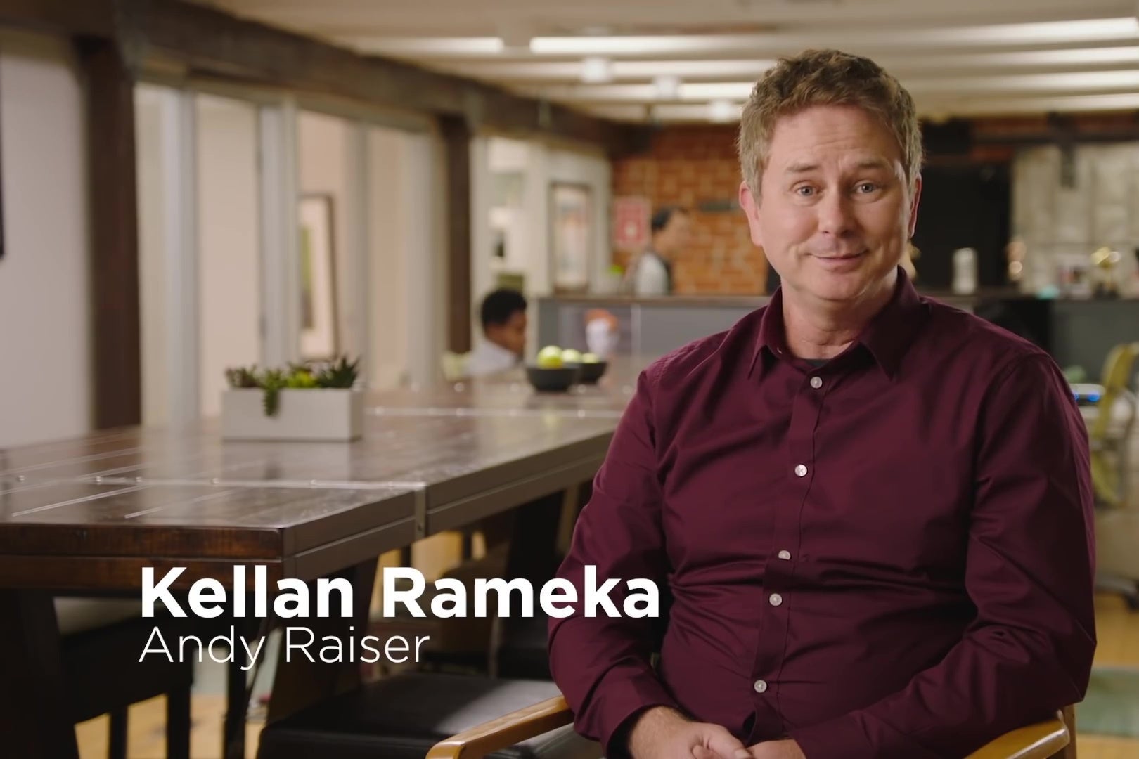 A man in a dark red shirt sits in an industrial office space, looking politely at the camera. Text identifies him as "Kellan Rameka: Andy Raiser."
