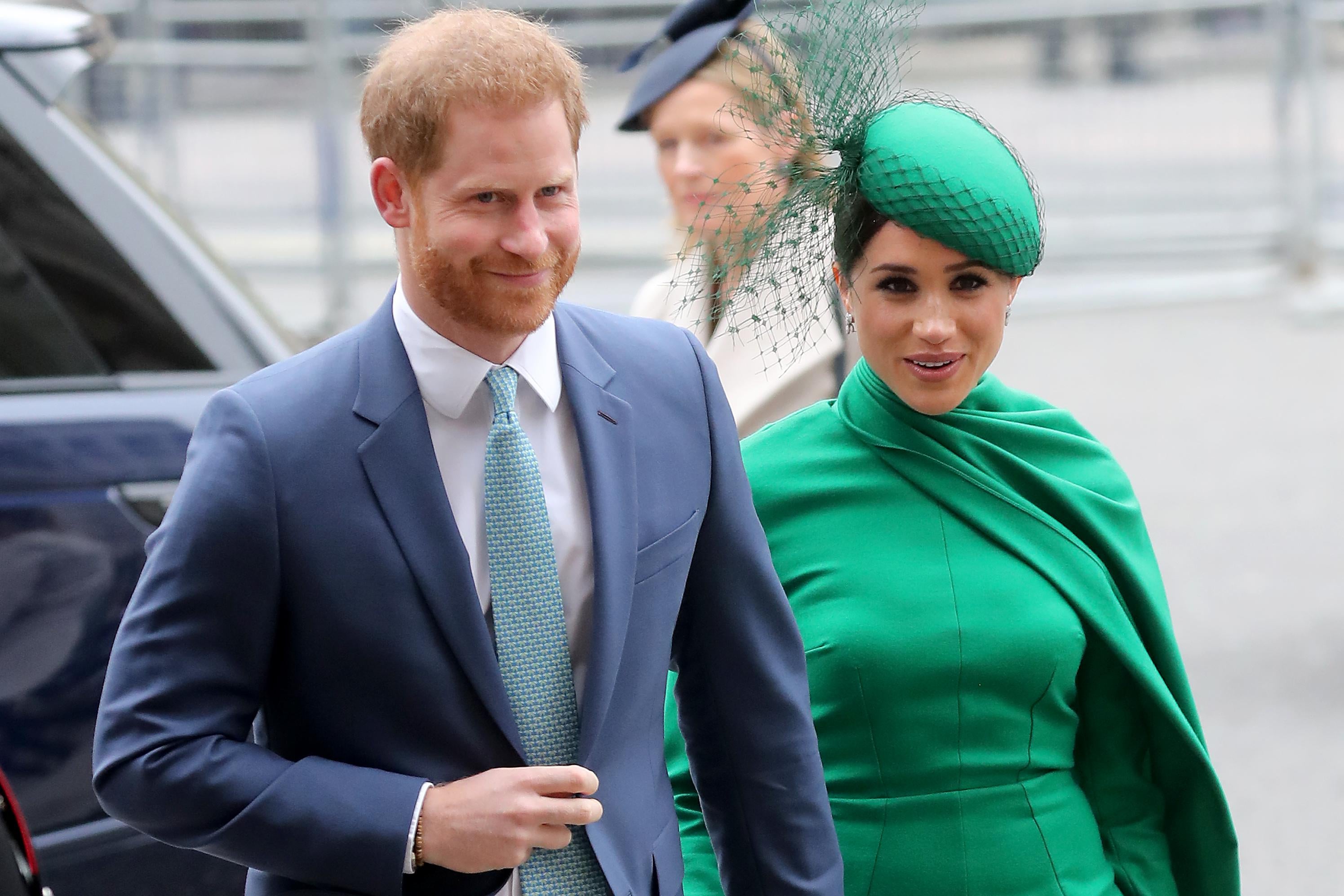 Prince Harry wearing a suit standing next to Meghan Markle in a green outfit with cape and green hat, seen from the waist up, in London.