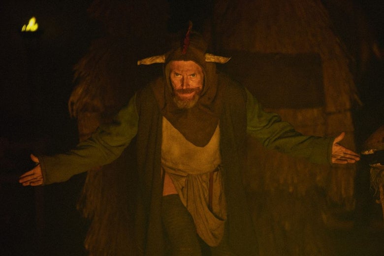 A man in a robe and hood stands in front of a hut with his arms outstretched.