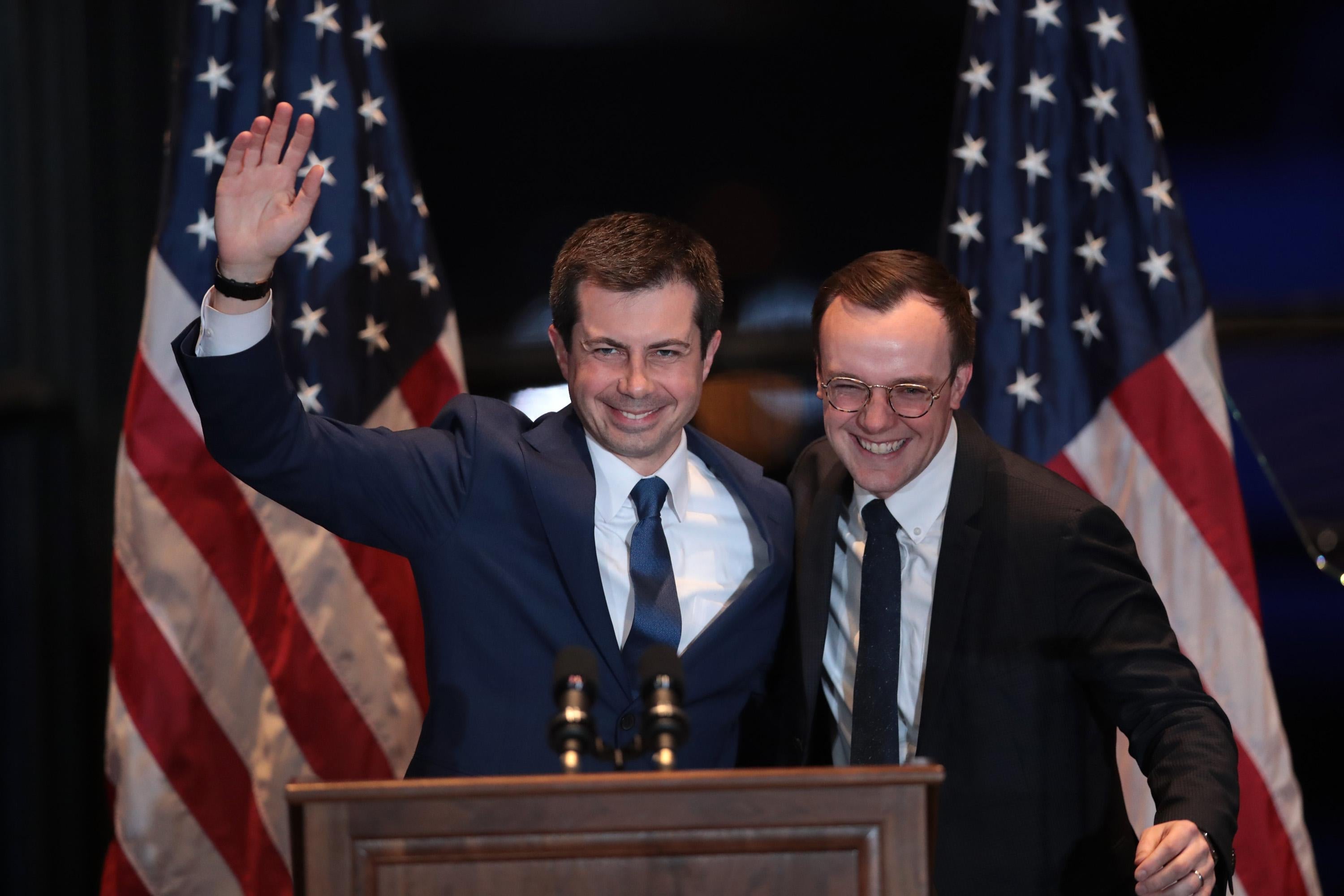 Pete and Chasten Buttigieg hugging and waving in front of American flags