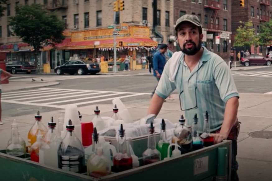 Lin Manuel Miranda, wearing an NYC cap and a cloth slung over his shoulder, pushes a cart with syrups on a New York City street.