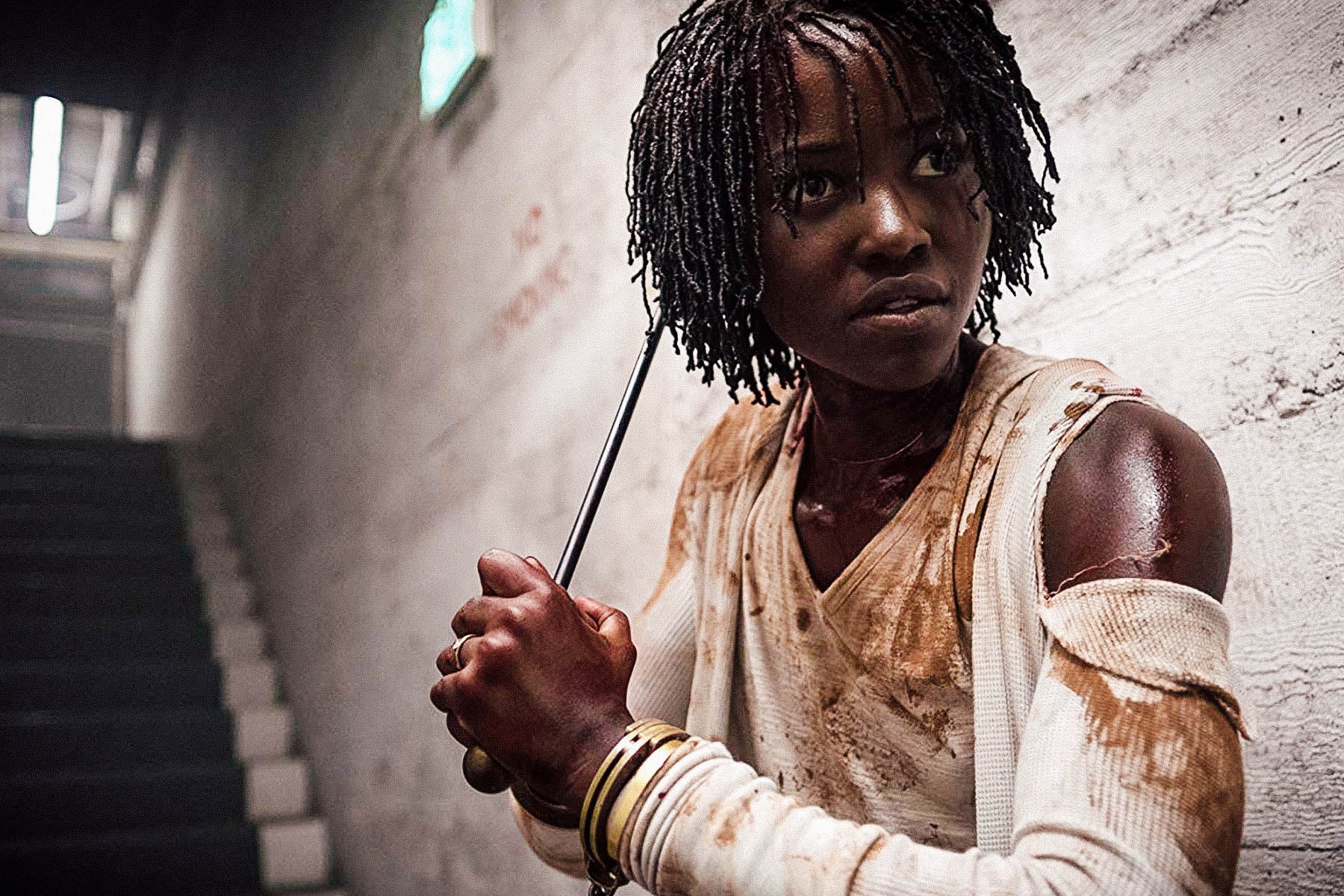 In a still from Us, Lupita Nyong’o stands at the foot of a set of stairs in a deserted hallway. She is wearing dirty clothing and brandishing a metal rod as a weapon.