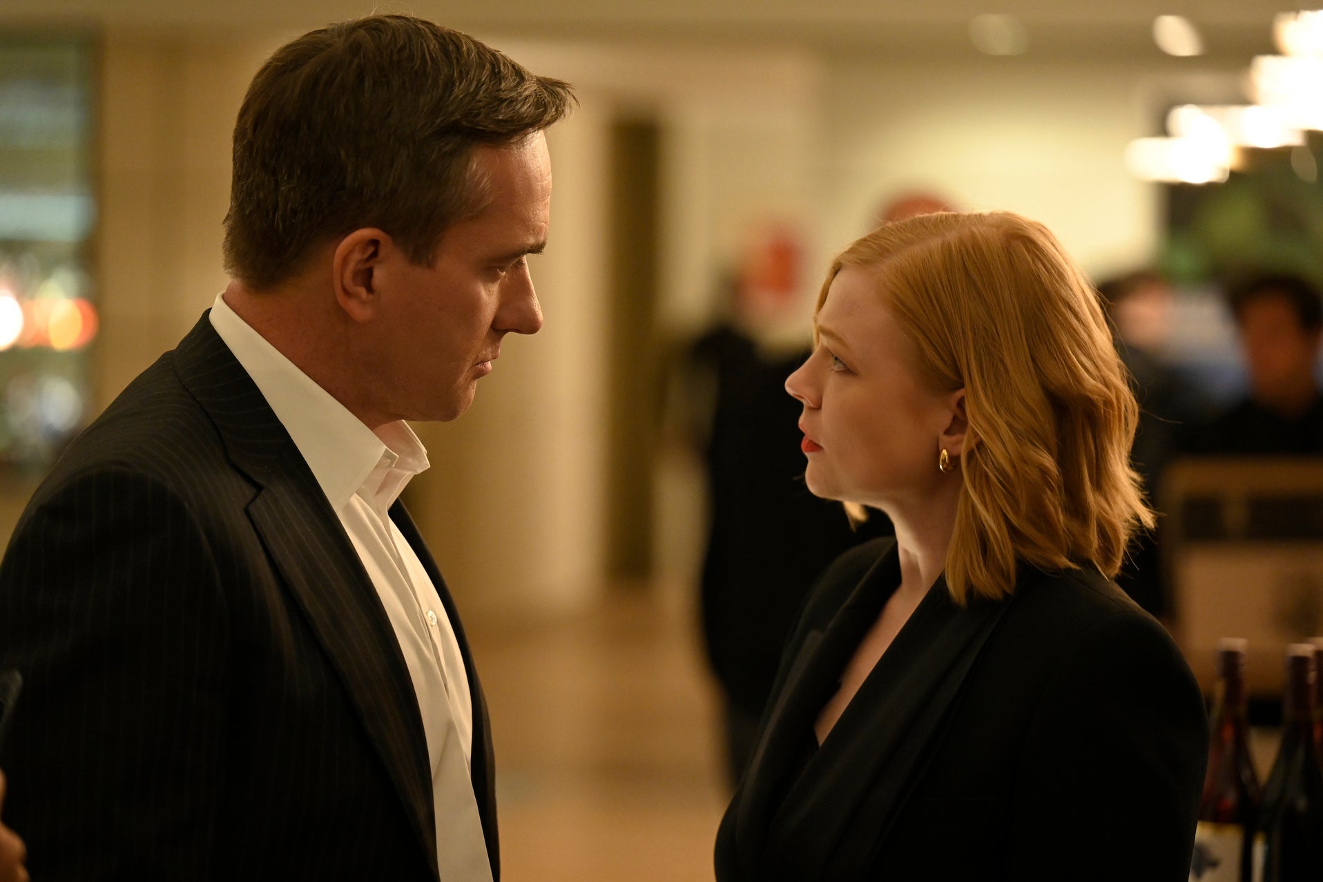 Tom Wambsgans (Matthew Macfadyen) and Shiv Roy (Sarah Snook) are dressed up in nearly matching black blazers, in a formal setting with wine bottles behind them, giving an intense and not entirely happy look.