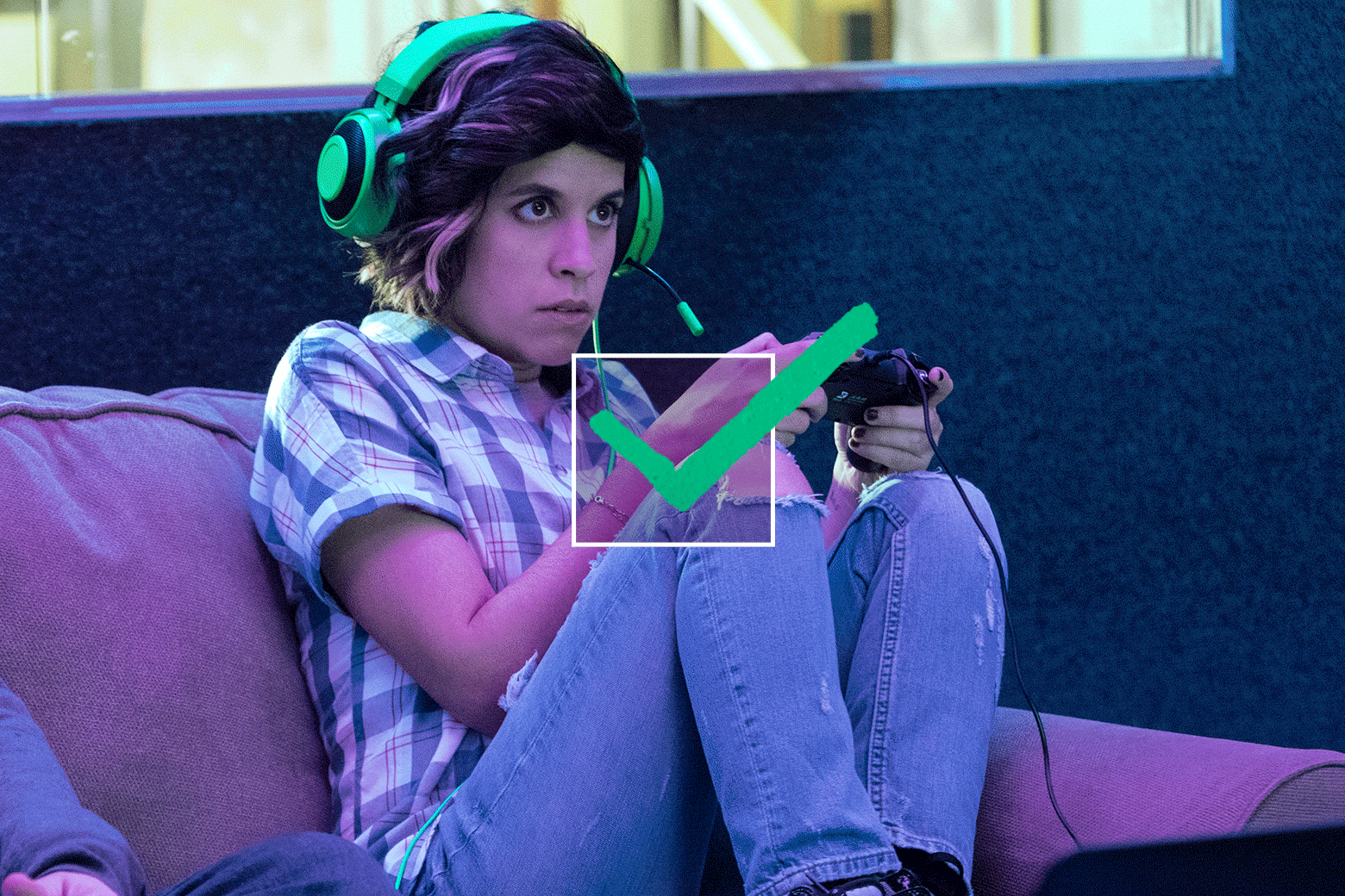 A woman sits on a couch playing a video game. There is a green checkmark animation over the photo.