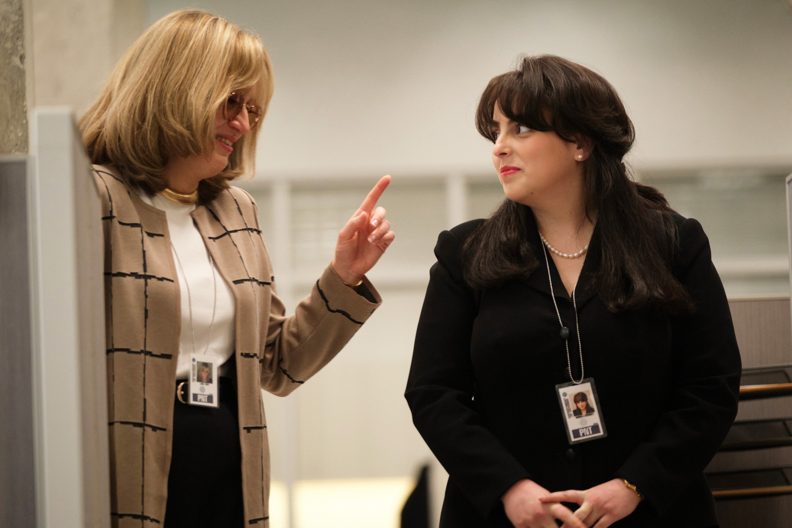 Sarah Paulson as Linda Tripp raises her left index finger at Beanie Feldstein as Monica Lewinsky as they stand together in an office, smiling collegially