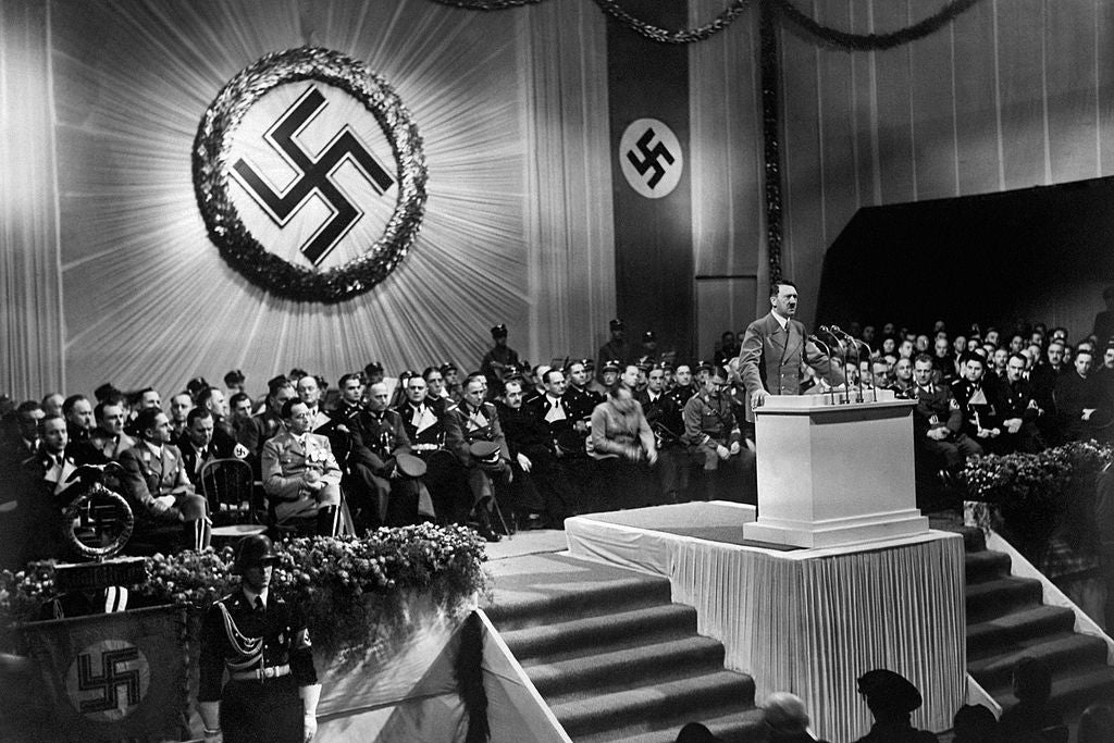 Hitler speaks from a crowded dais in front of a large Nazi insignia.