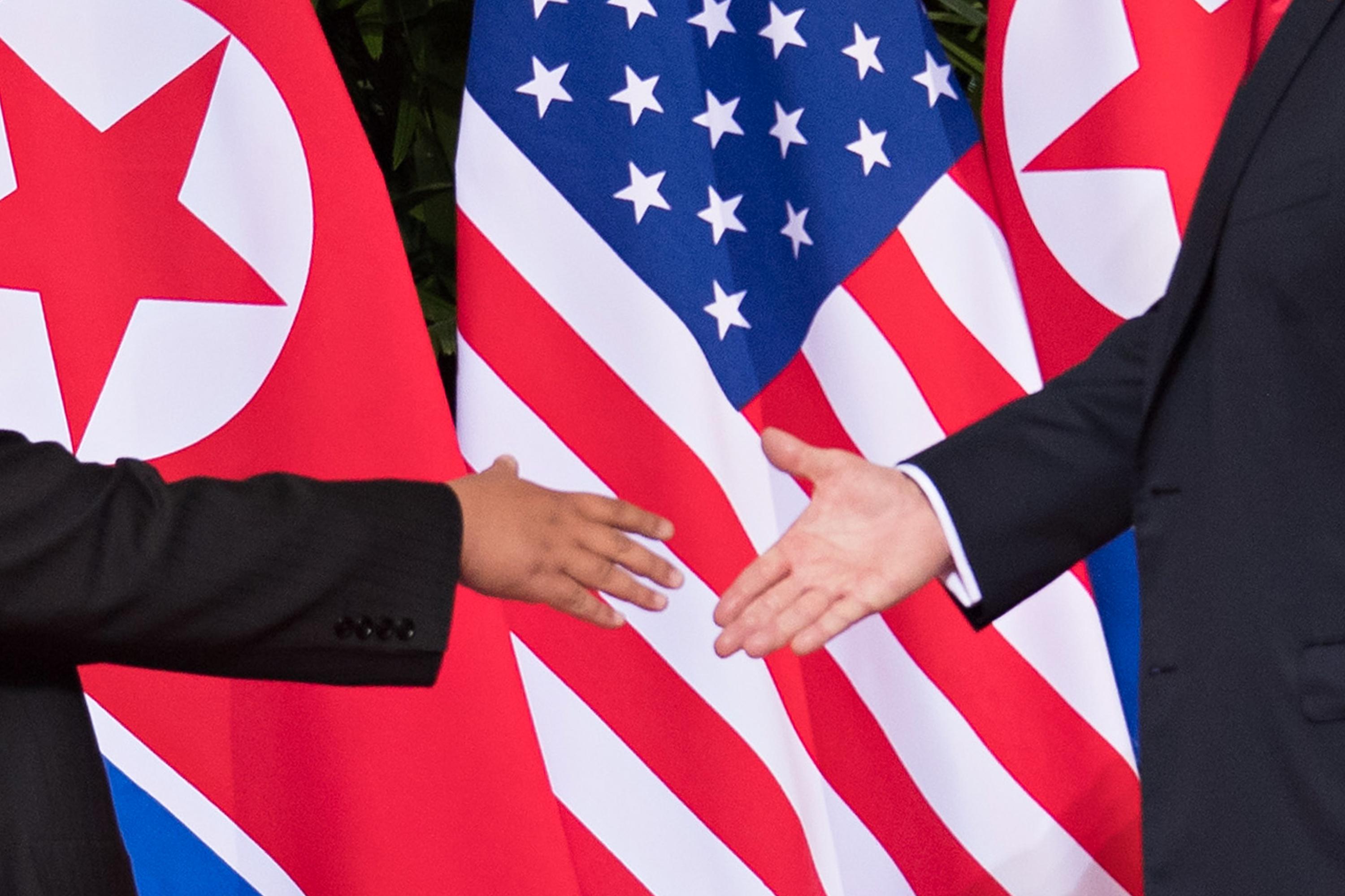 Kim Jong-un and Donald Trump shake hands in front of North Korean and U.S. flags.