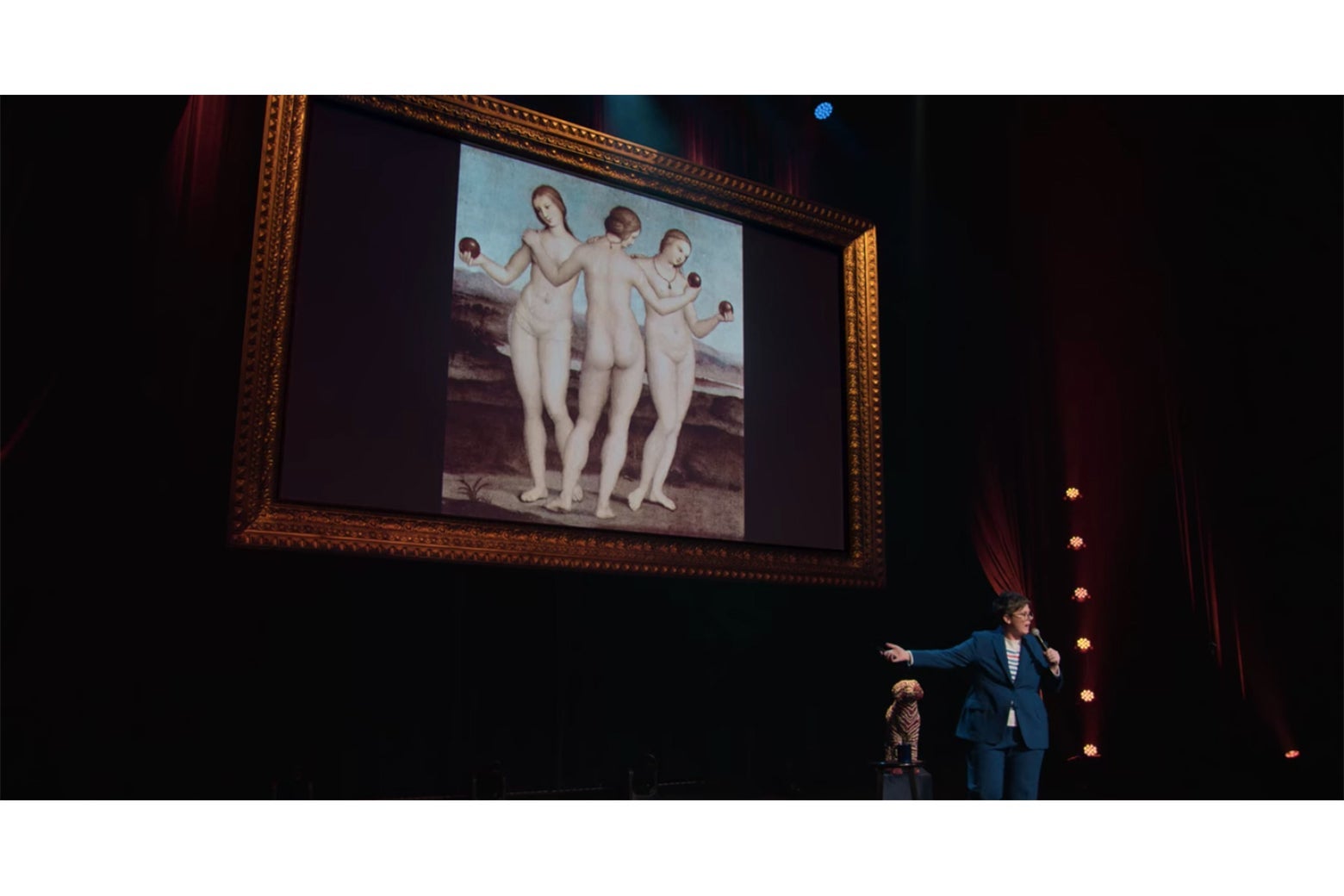 Hannah Gadsby standing in front of an image of Raphael's Three Graces.