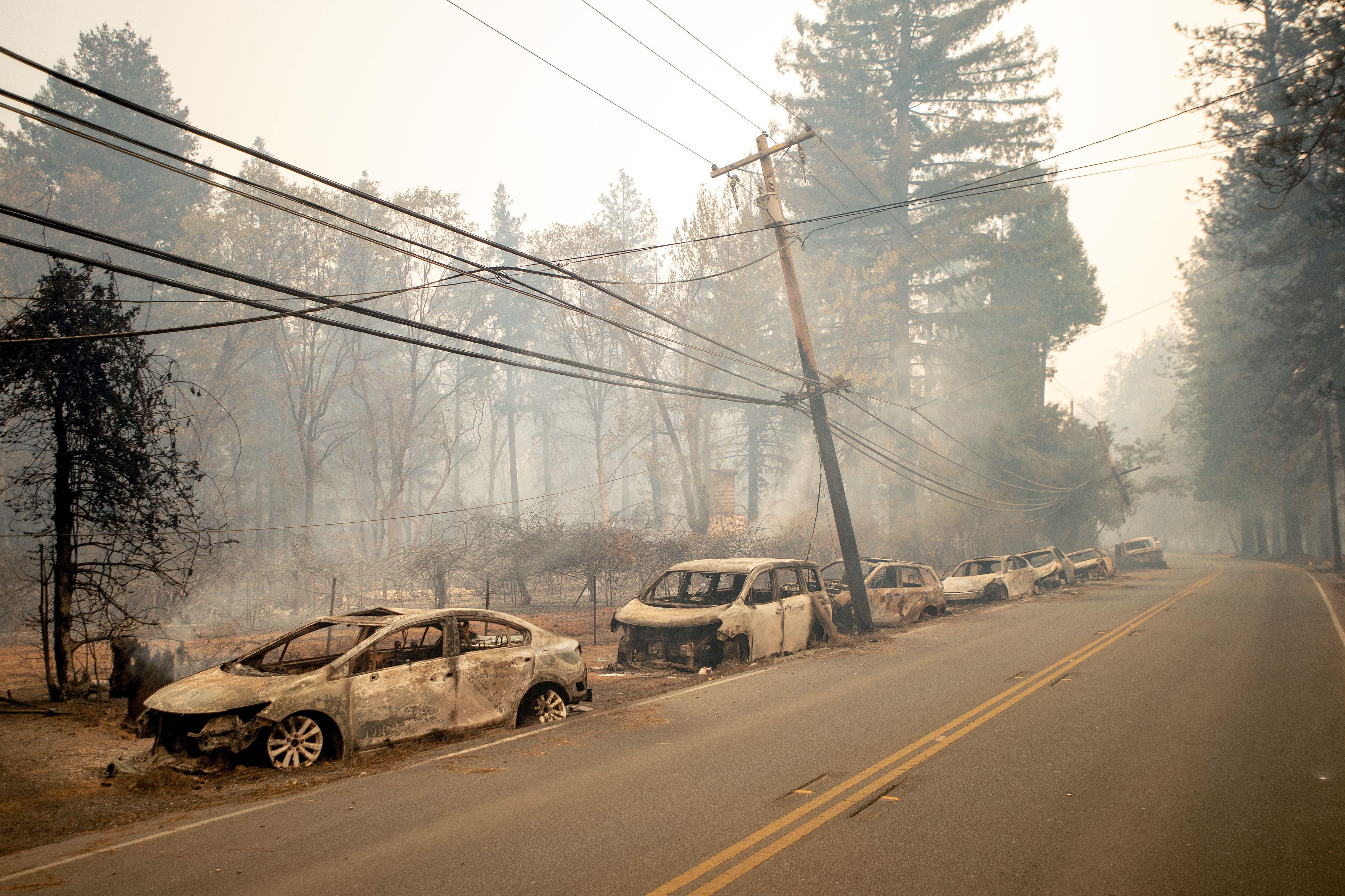 Abandoned and charred vehicles sit on the side of a road. An electrical pole leans dangerously. The air is hazy.