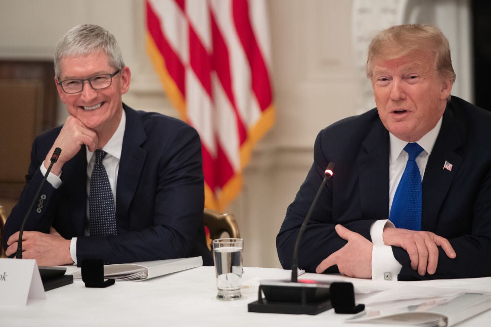 Tim Cook and Donald Trump, wearing suits, sit at a conference table in front of an American flag.