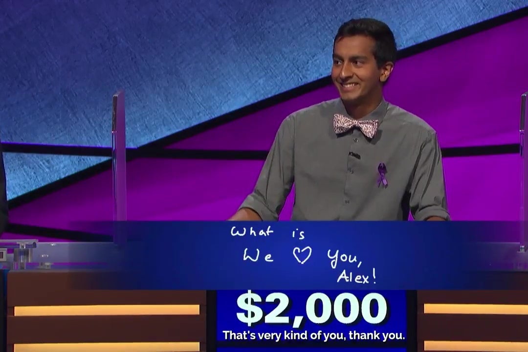 Jeopardy contestant Dhruv Guar stands behind his Jeopardy! podium, smiling; his Final Jeopardy answer, "What is, We ♡ You, Alex!” is projected above his podium.  