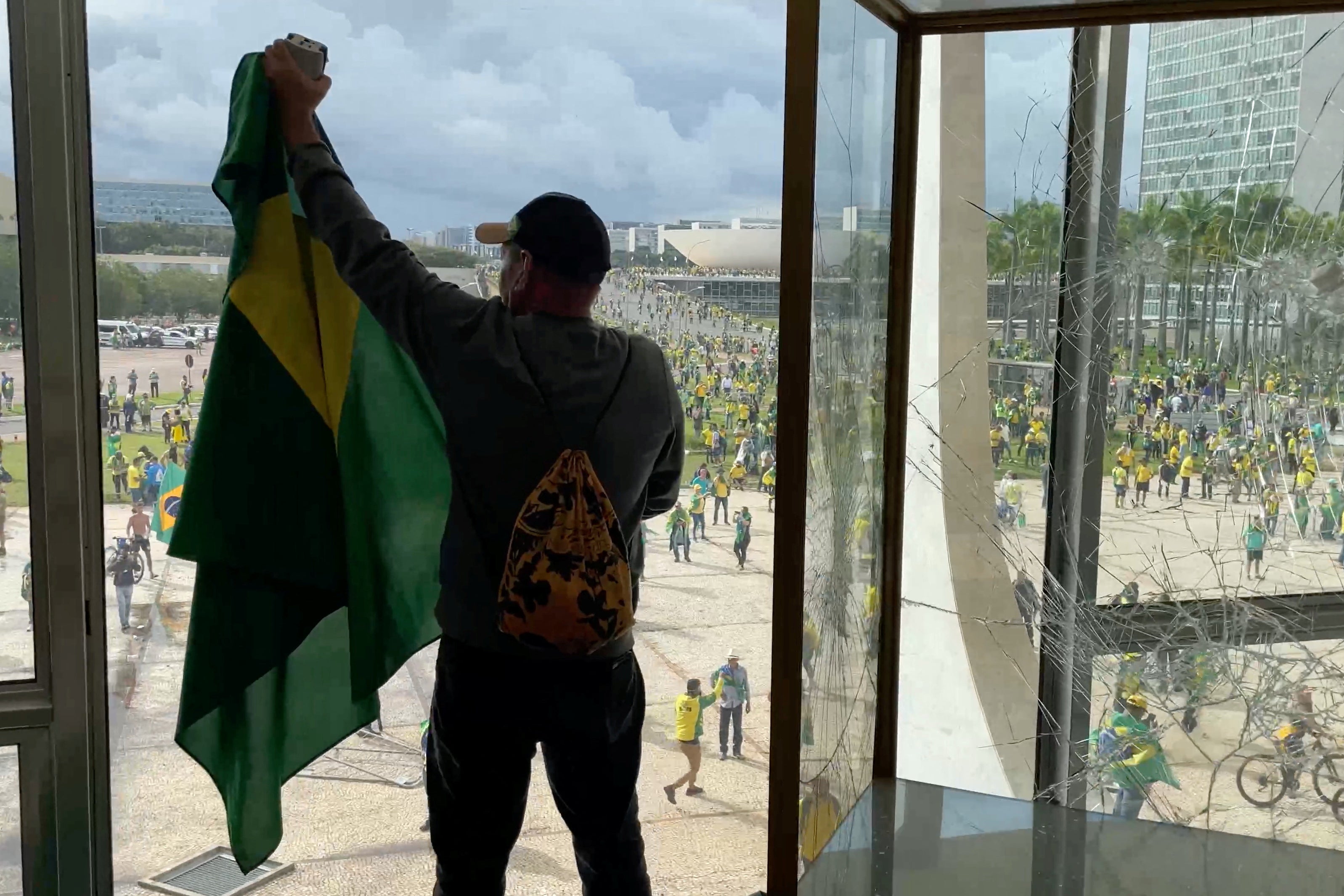 A man holds up a Brazilian flag from inside a government building with shattered windows. A crowd of green-and-yellow clad protesters can be seen outside.