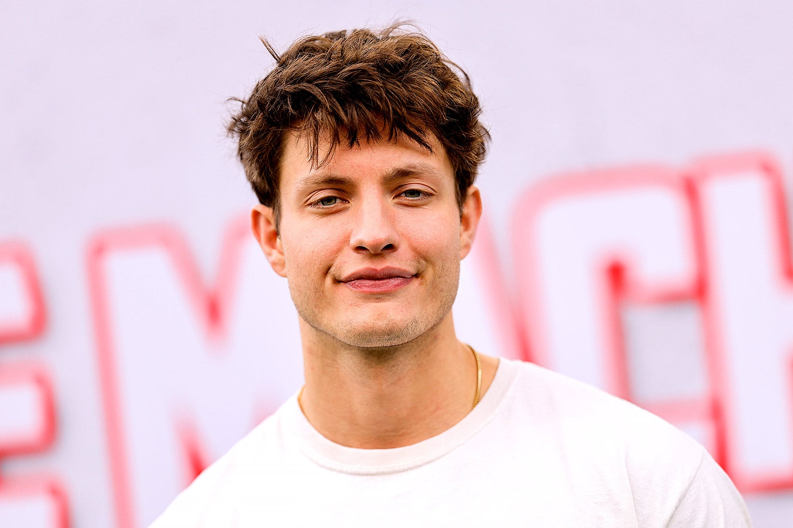 A brown-haired man in front of a pink background wears a white T-shirt and smiles slightly.