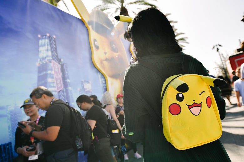 A person in a black hood wears a yellow backpack with a smiling face on it. They are facing a crowd of people in black t-shirts who are looking at their phones. 