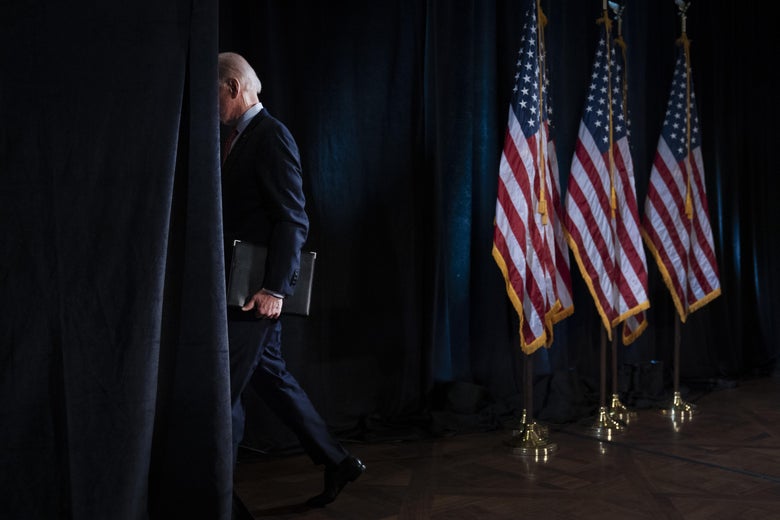 Joe Biden walks through a gap in a wall of curtains, carrying a binder, next to three American flags.