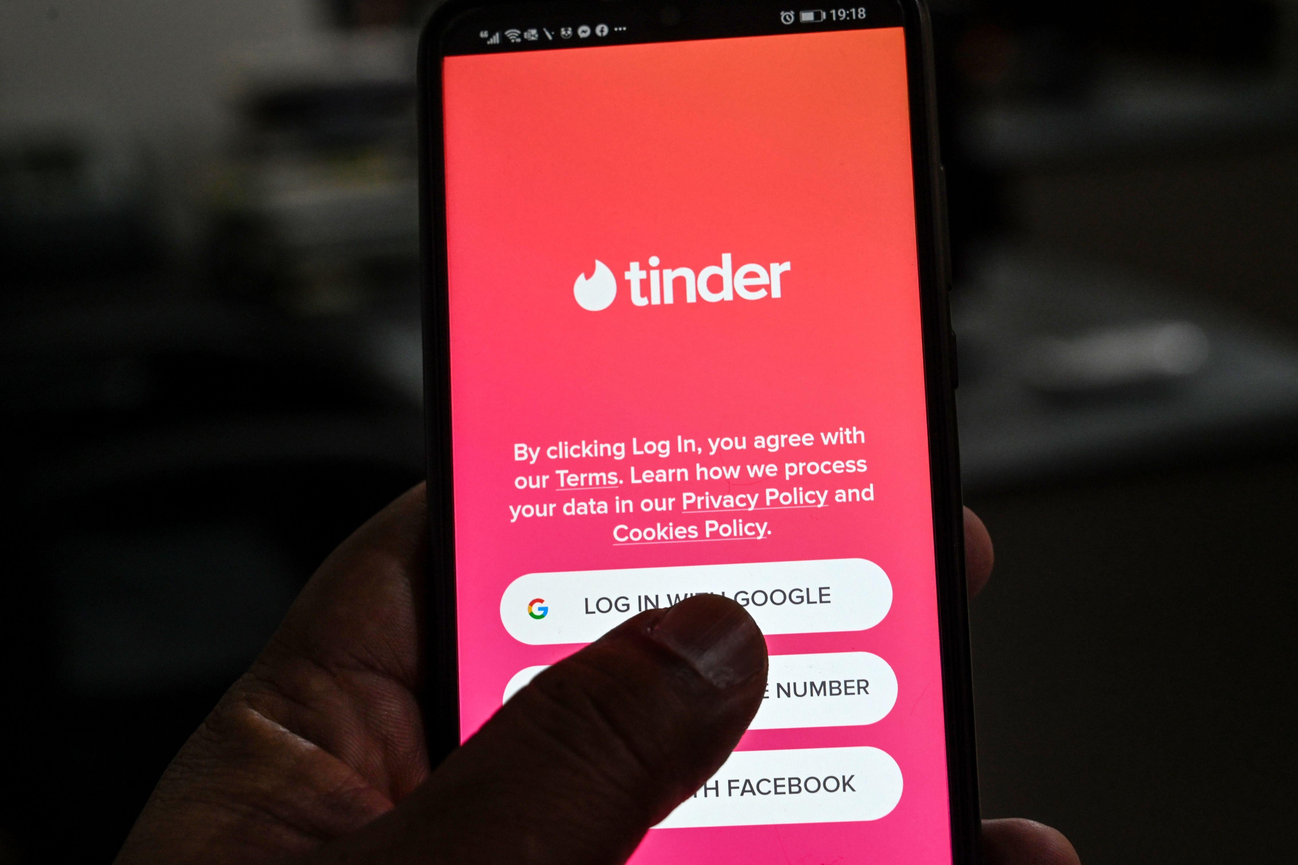 A close-up of a person holding their phone, with their thumb on the screen. They are on the log-in page for Tinder, which has a pink background and the name of the app in white. 