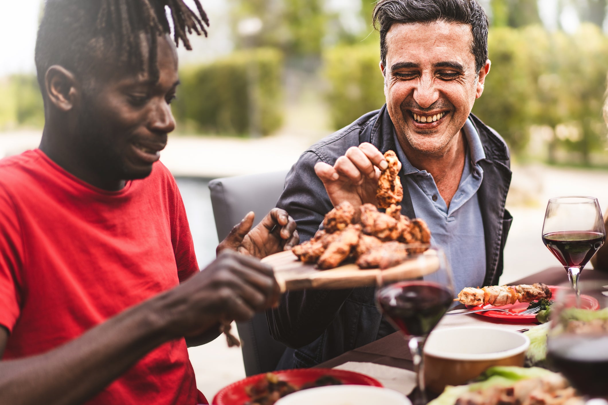 A man in a red shirt hands a tray of chicken wings to another man at an outdoor table lined with wine and food. Both men are smiling.