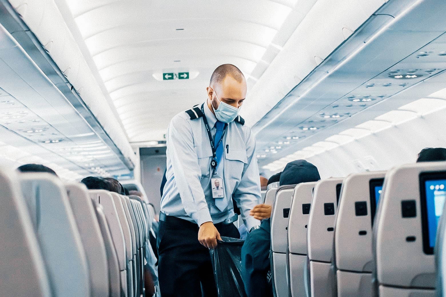 A flight attendant wearing a surgical mask walks down the center aisle of a plane, collecting trash from passengers