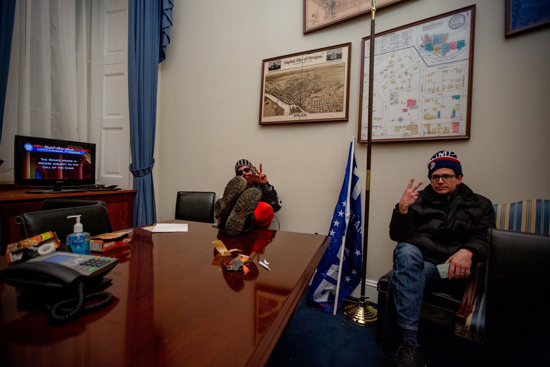 Two men sit at a table in the U.S. Capitol, one flashing a peace sign.