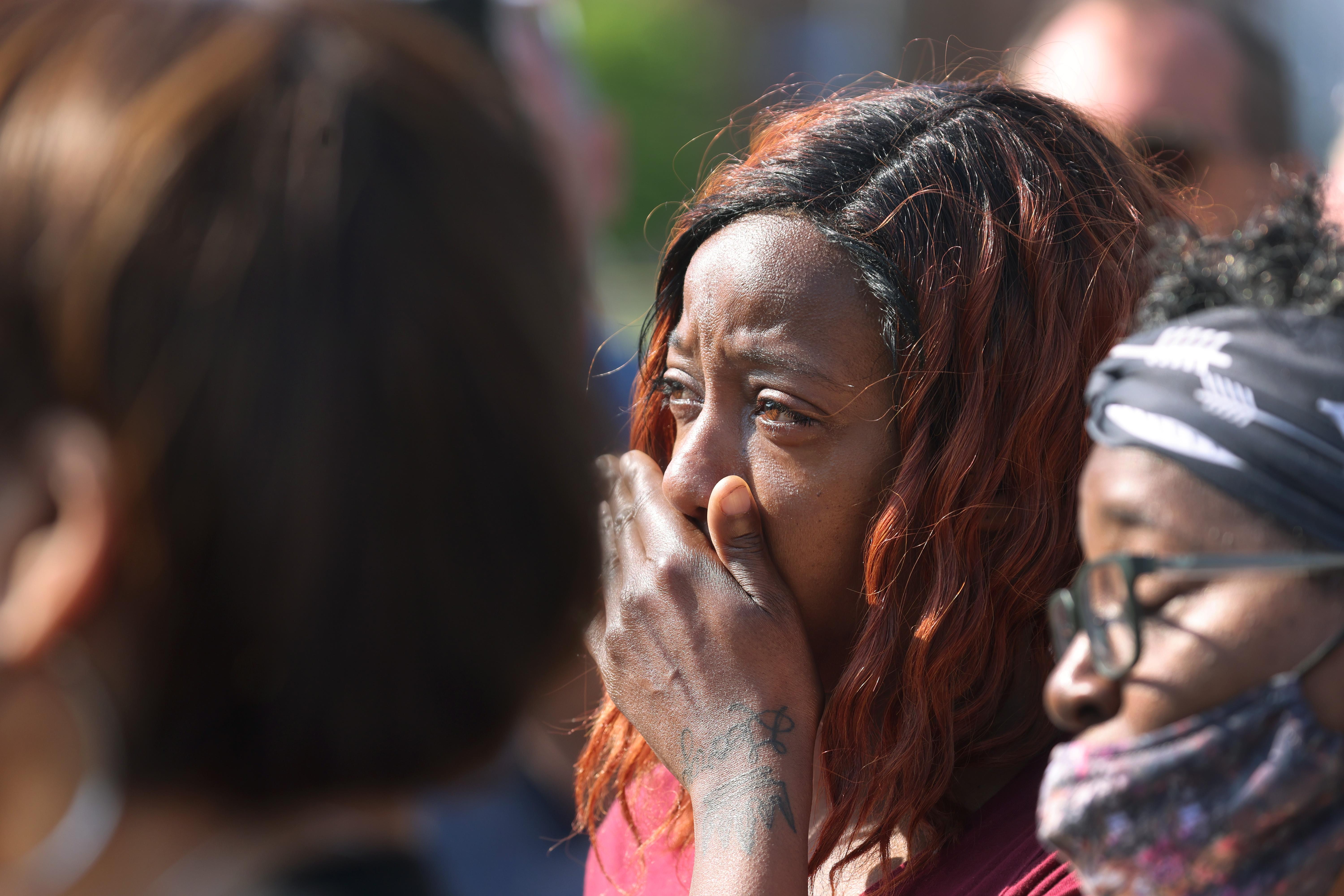 A woman stands in a crowd of mourners holding her hand to her mouth in horror.