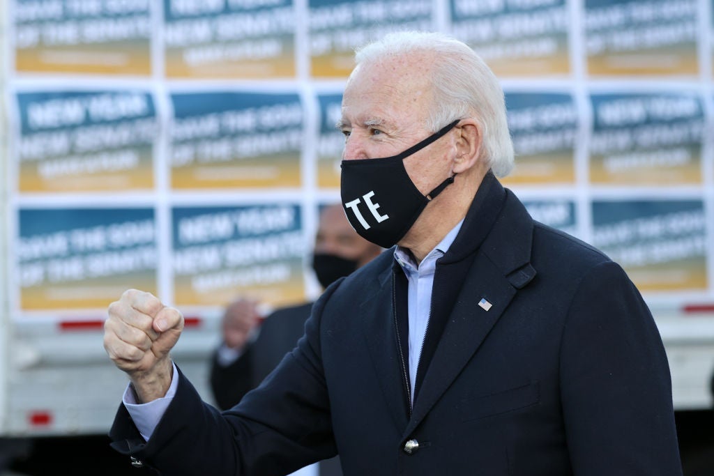 Biden, wearing a black mask and a coat, pumps his fist on a stage.