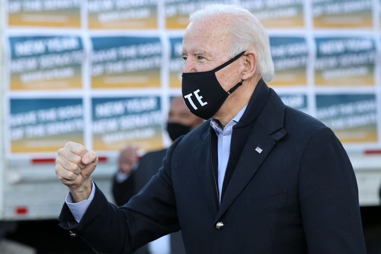 Biden, wearing a black mask and a coat, pumps his fist on a stage.