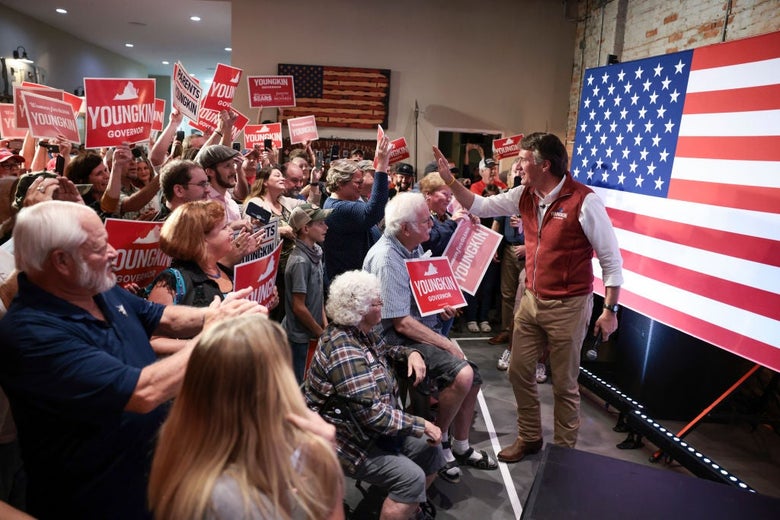 Youngkin, wearing a red vest and standing in front of an American flag, waves to a tightly packed-in group of supporters holding red signs.