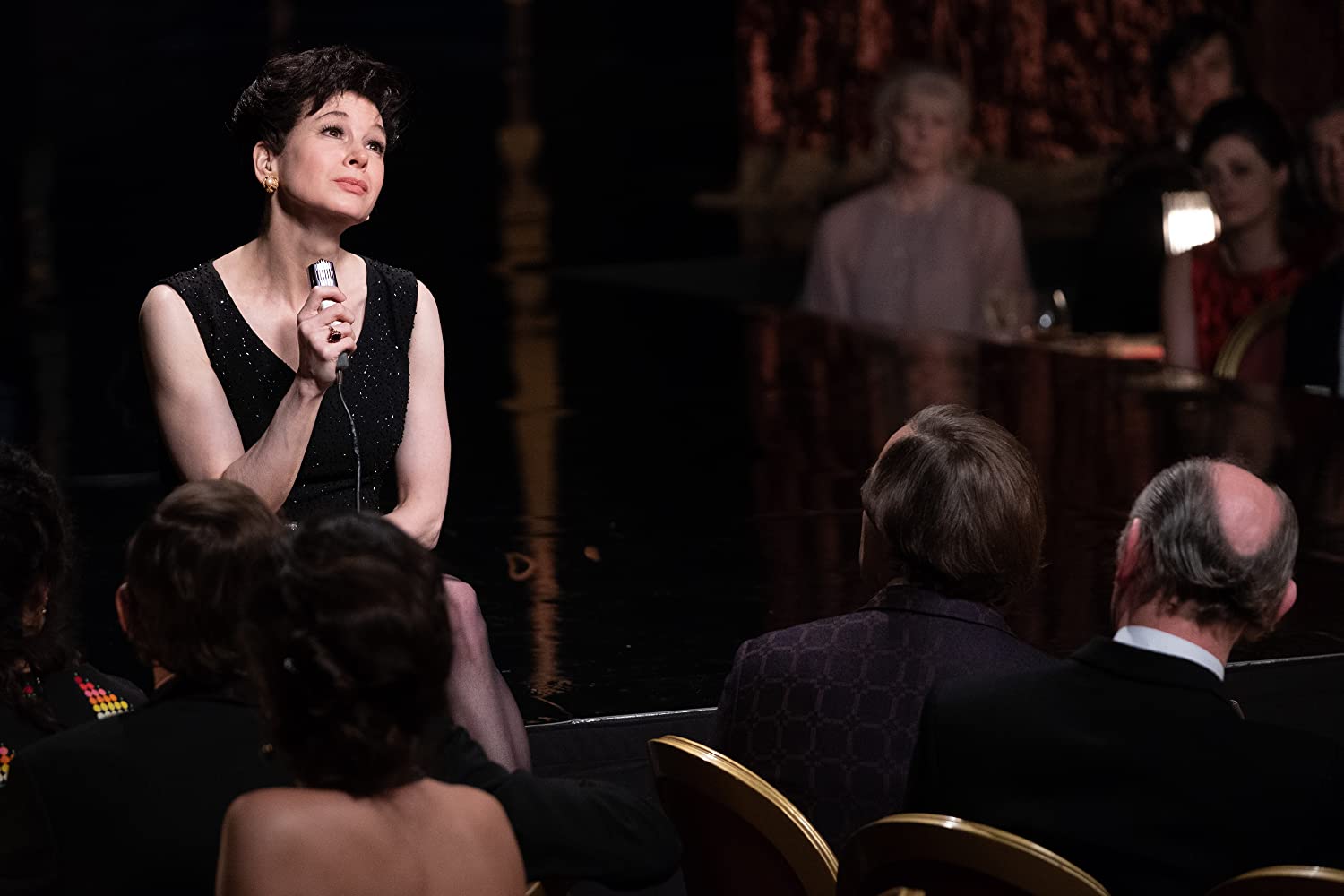 Renée Zellweger sits with a microphone in front of onlookers at tables.
