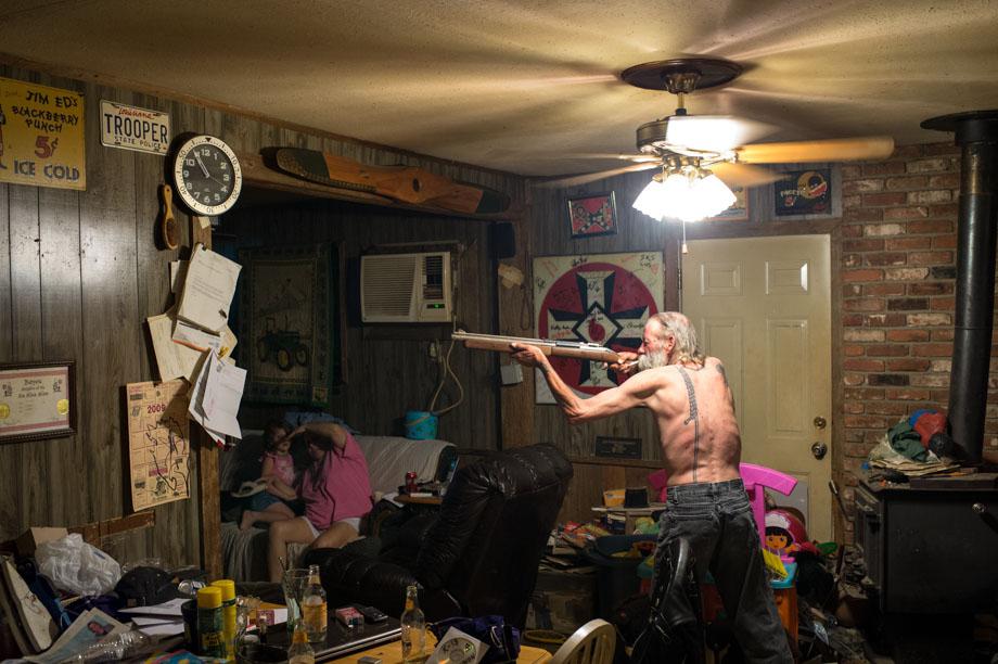 Carl, an Imperial Wizard of a southern-based Ku Klux Klan realm, takes aim with a pellet gun at a large cockroach (on the piece of paper just below the clock) while his wife and goddaughter try to avoid getting struck by a possible ricochet.