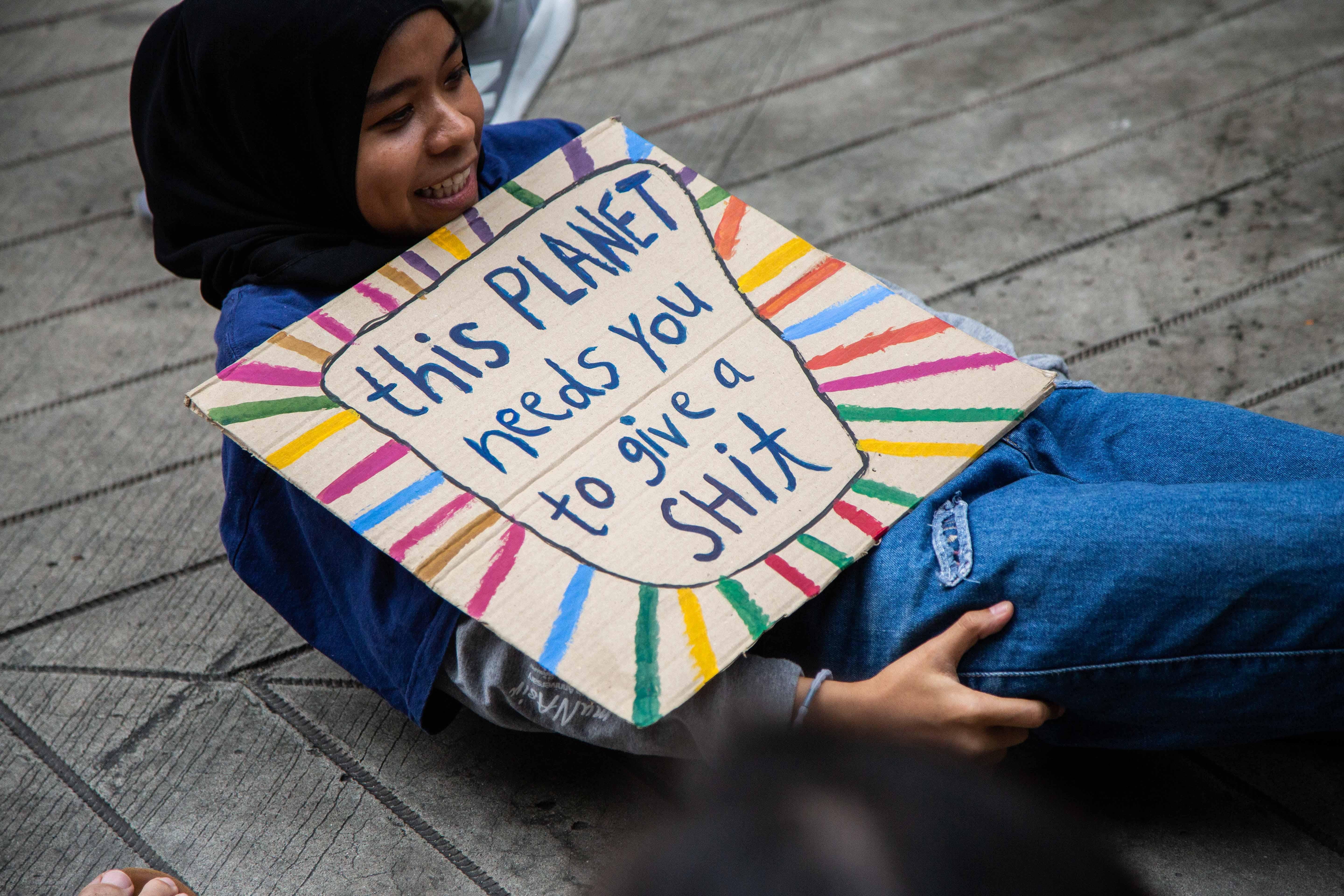 A girl holds a sign that says "this planet needs you to give a shit."