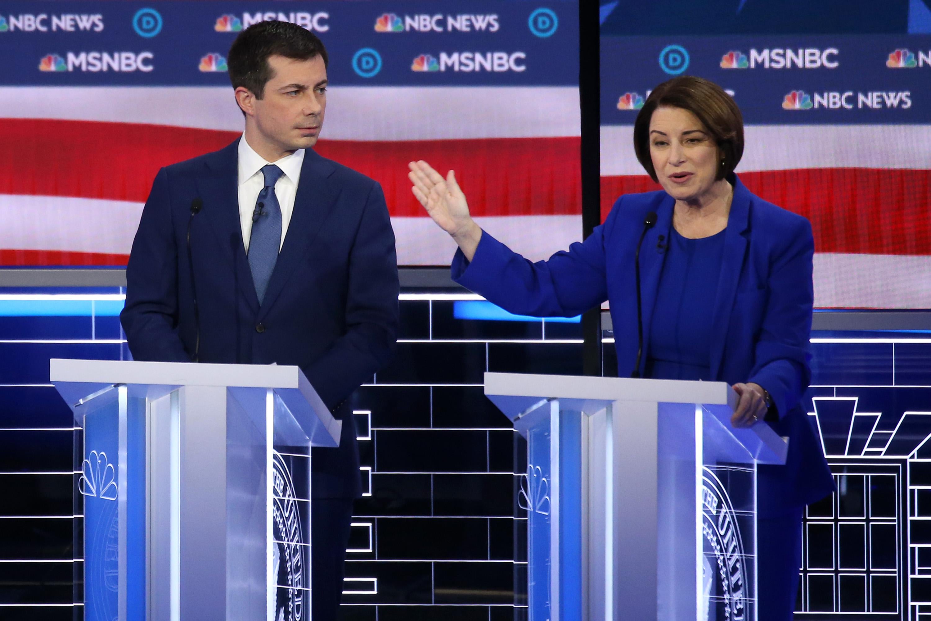 Pete Buttigieg and Amy Klobuchar seen on a debate stage. Klobuchar gestures toward Buttigieg.