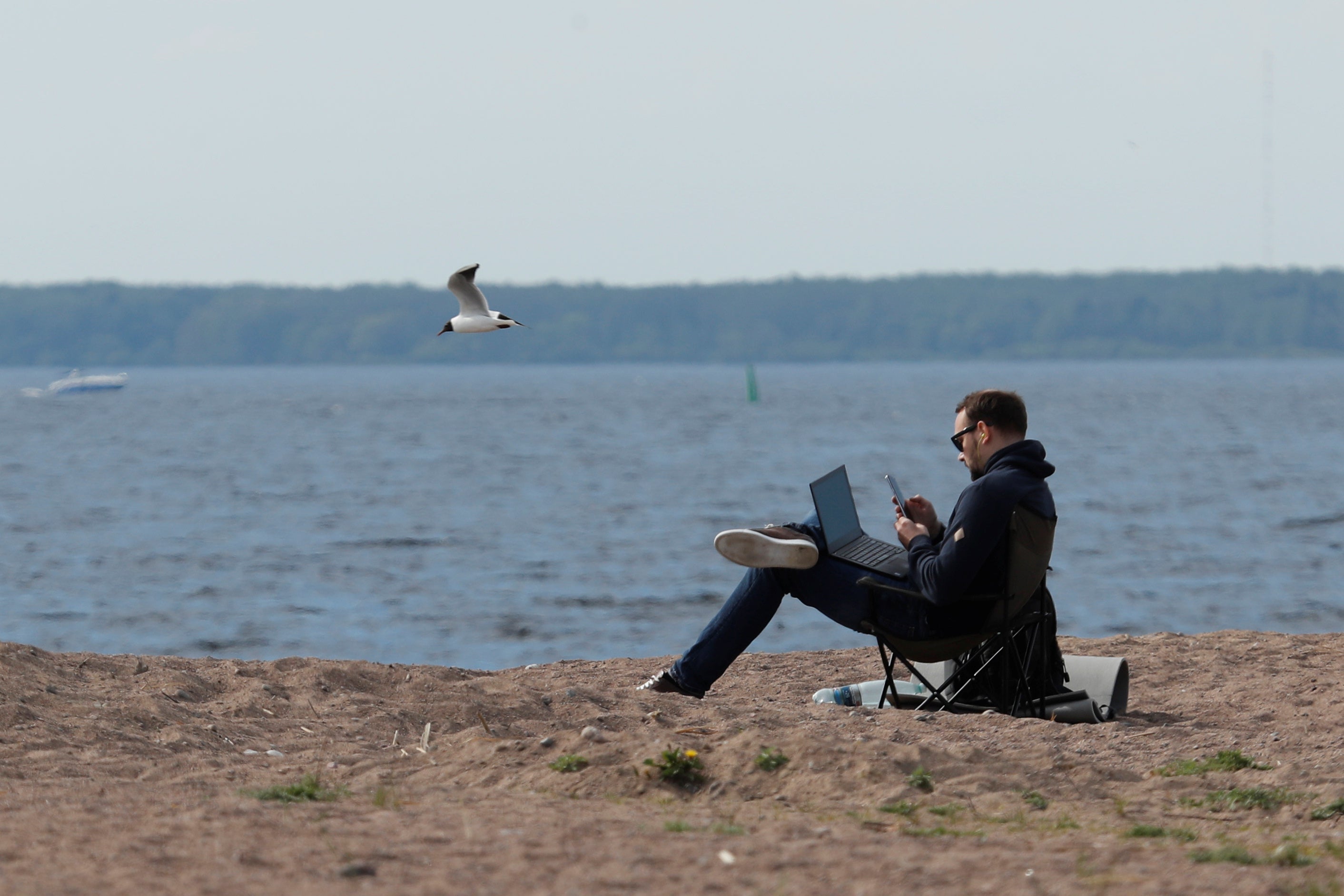 A man uses his smartphone and has a laptop in his lap as he sits in a camping chair on a beach