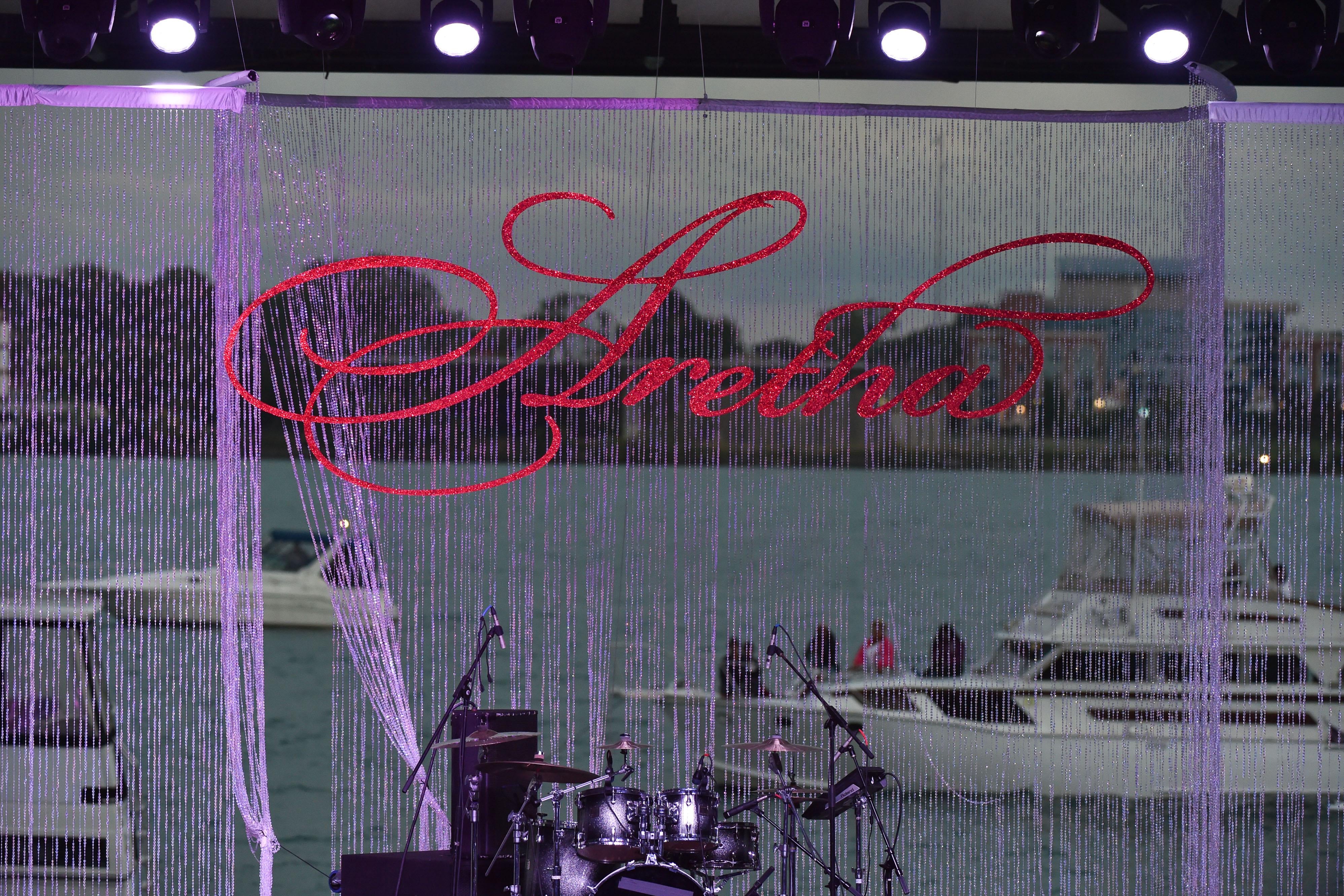A drum set on an empty stage. In the background, an image of boats on a river, streaked with purple string, and the word Aretha in red script.
