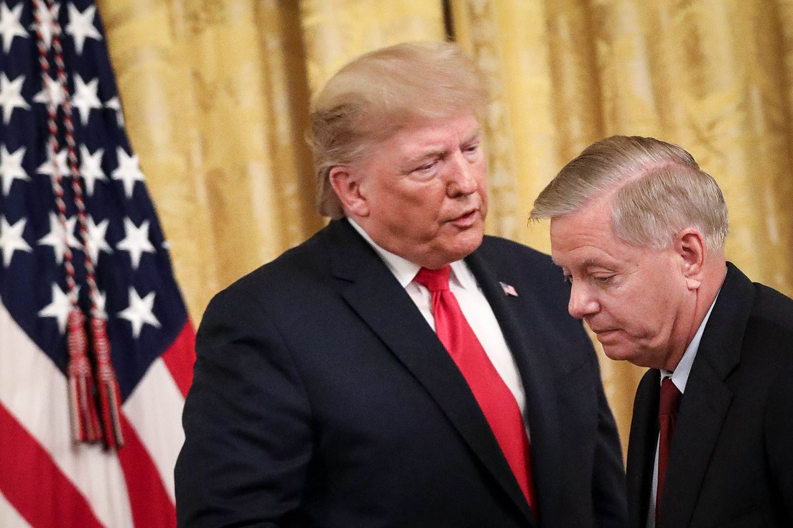 Donald Trump opens his mouth as if to talk as Lindsey Graham walks by. There is a gold curtain and an American flag behind them.