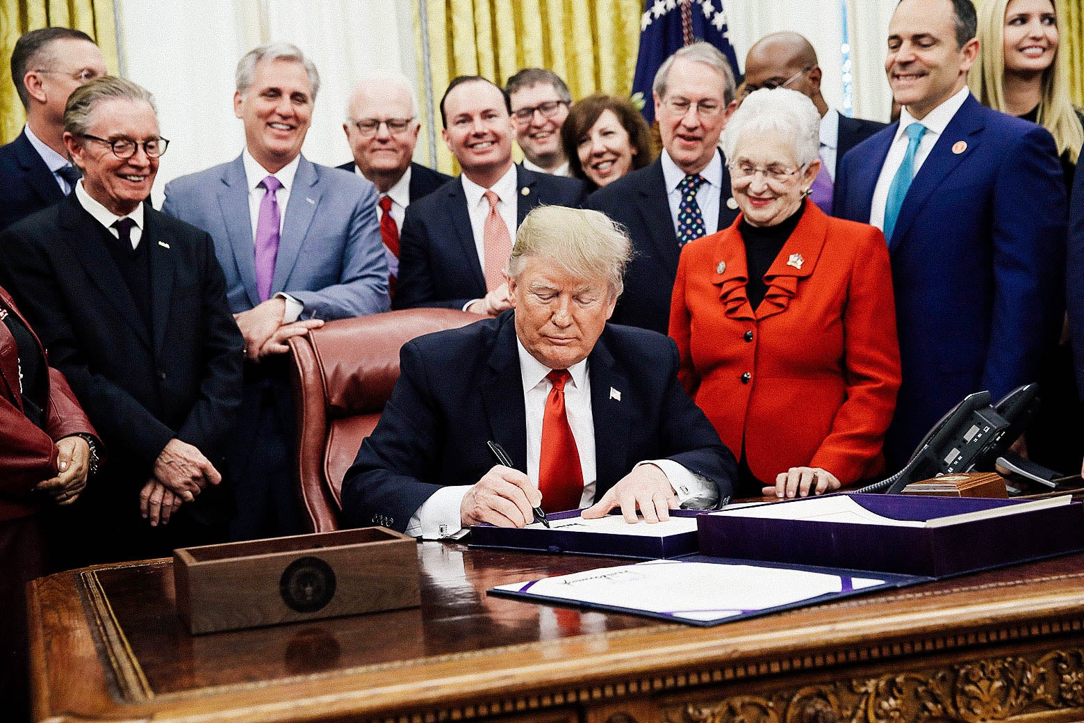 People surround Donald Trump, who's seated at his desk in the Oval Office signing a document.