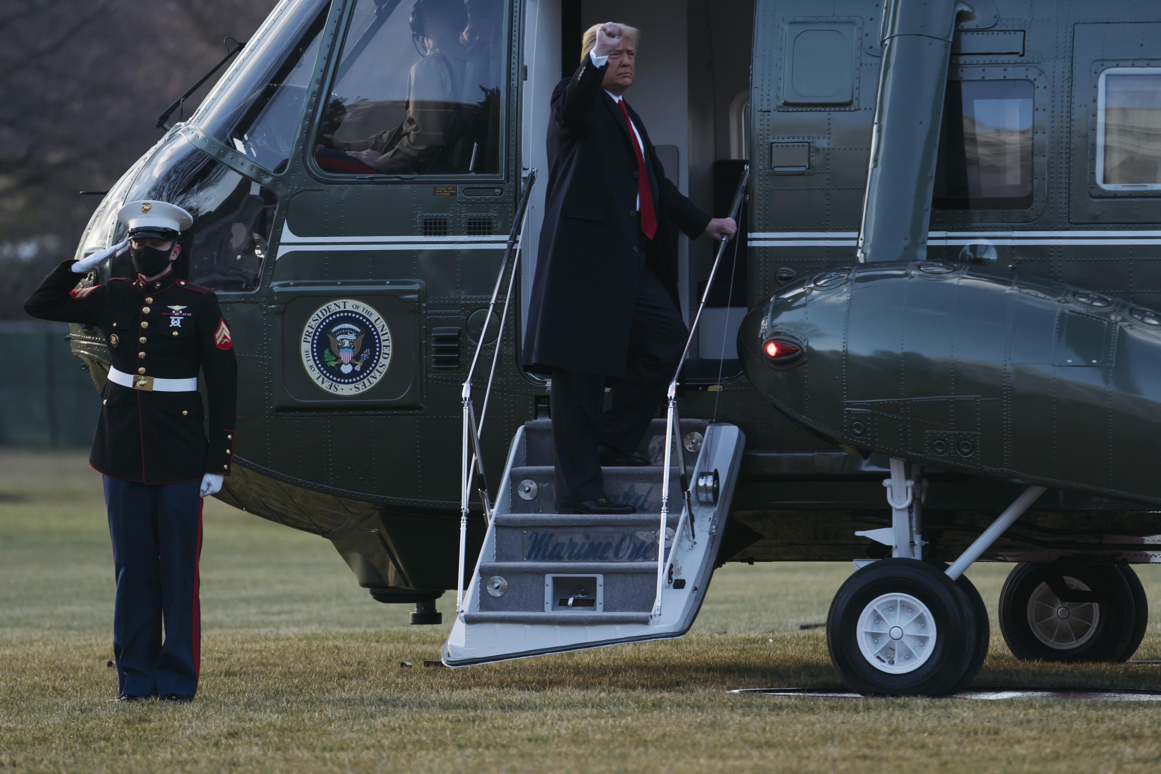 Donald Trump raises his fist to an audience off-screen as he stands on the stairs to Marine One. A masked Marine stands next to the helicopter, saluting.