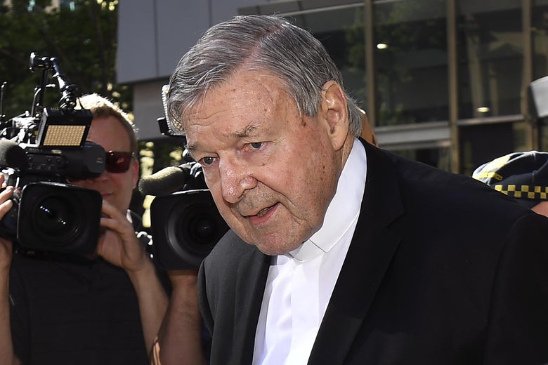 Pell, with his head bent downward, walks through a crowd of reporters. Behind him, a camera is trained on him.