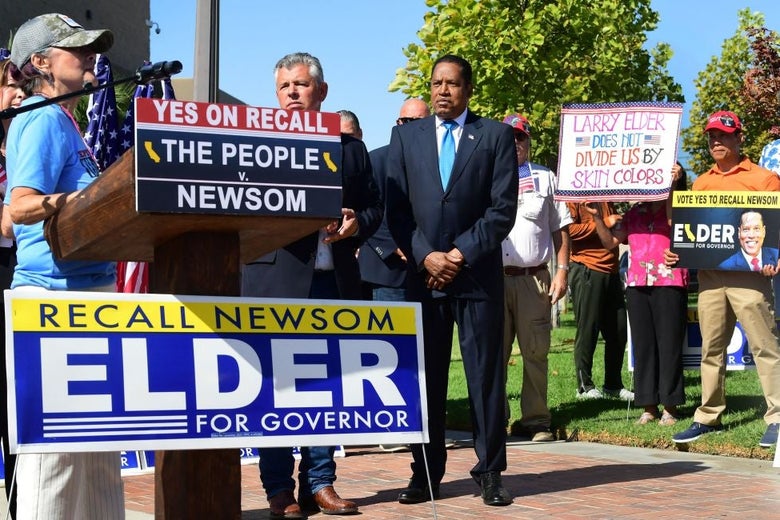 Elder, wearing a suit with a light blue tie, stands in front of a small group of supporters while waiting to be introduced to speak at a lectern in what appears to be a public park.