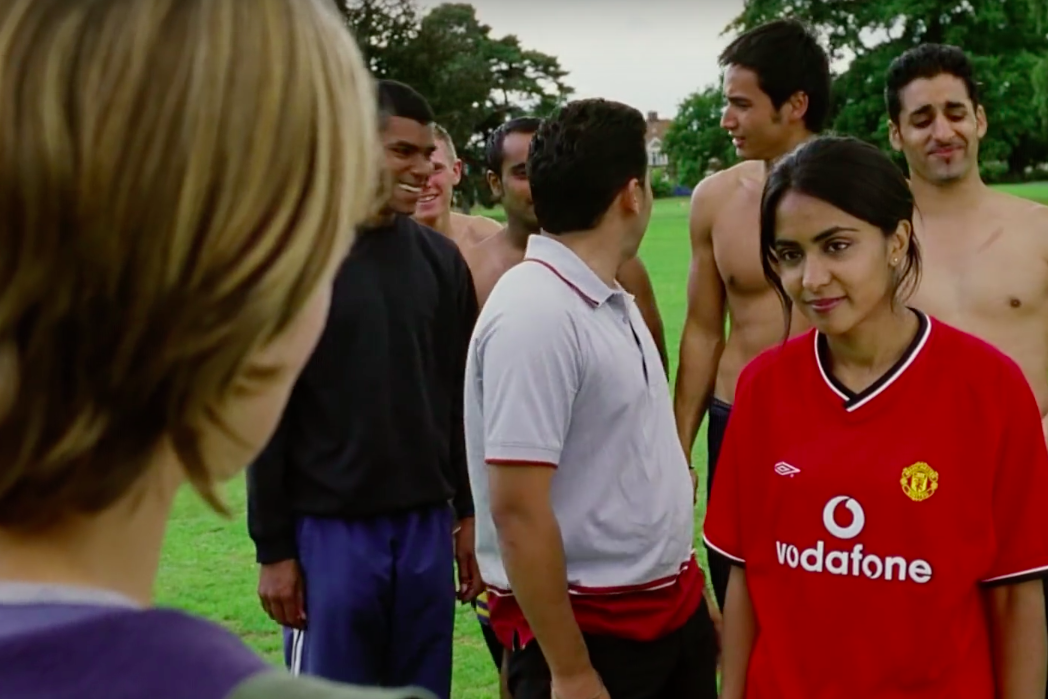 Parminder Nagra, wearing a red jersey, stands on a soccer field in front of a group of men, some of them shirtless.